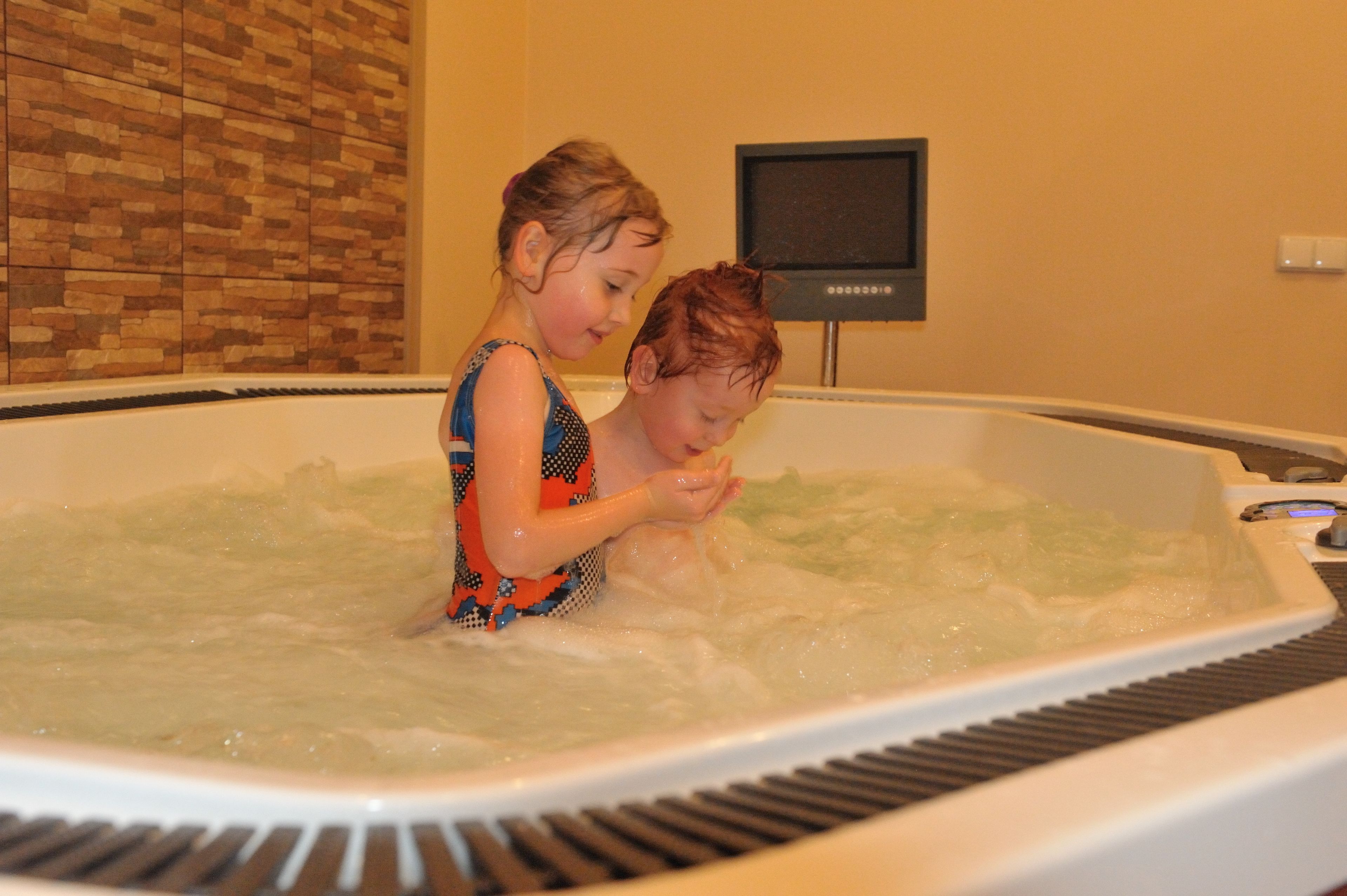 Dos niños en un jacuzzi con una pared de azulejos y un televisor al fondo.