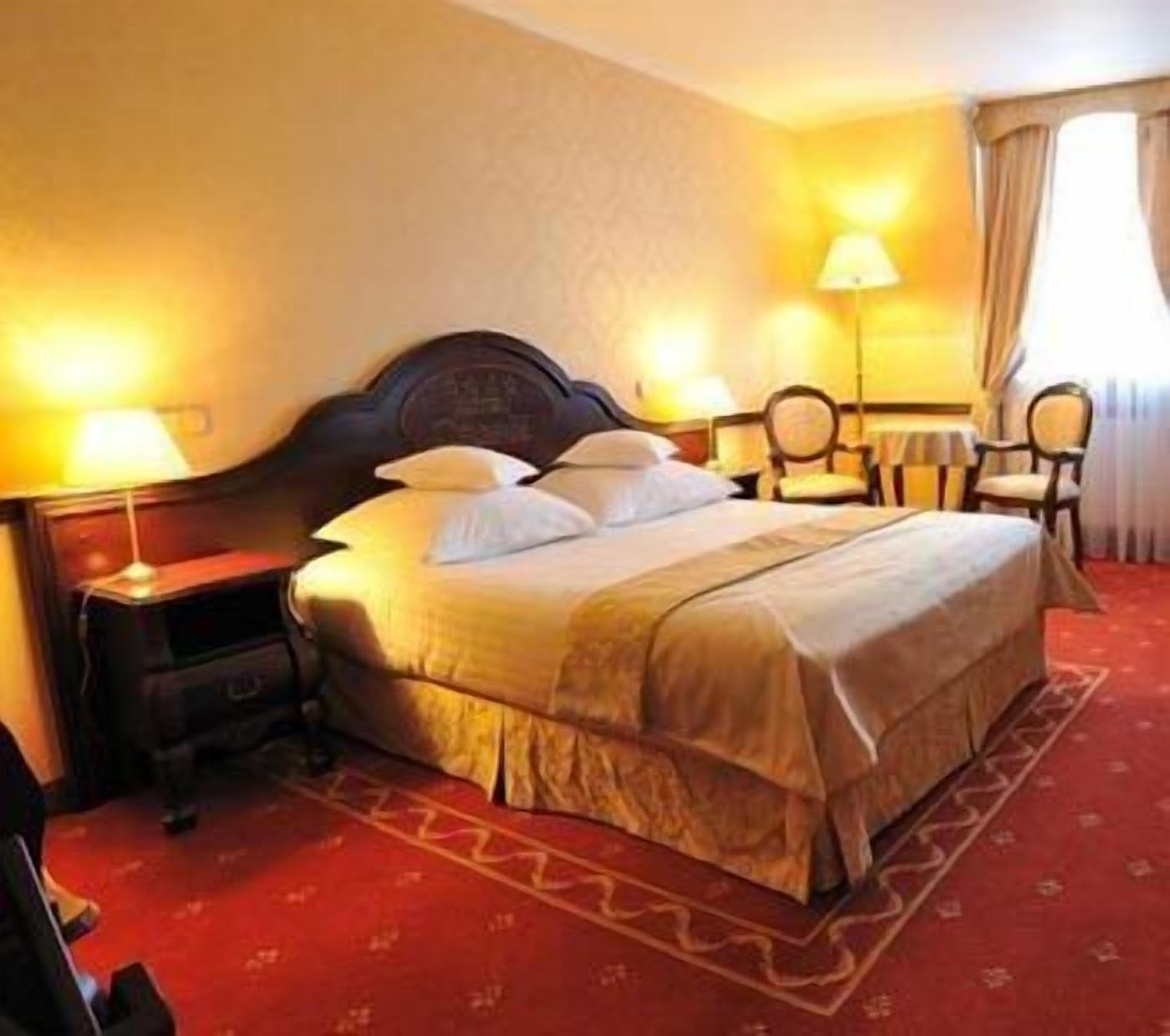 Habitación de hotel con una cama grande, dos mesitas de noche con lámparas, una pequeña mesa con sillas y un ventanal con cortinas.
