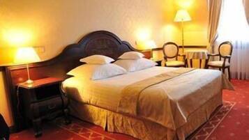 Habitación de hotel con una cama grande, dos mesitas de noche con lámparas, una pequeña mesa con sillas y un ventanal con cortinas.