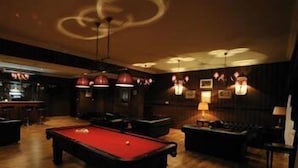 A billiards table in a dimly lit room with wooden floors and dark walls. There are bar stools and a bar in the background. - Hotel Chopin (Sochaczew)