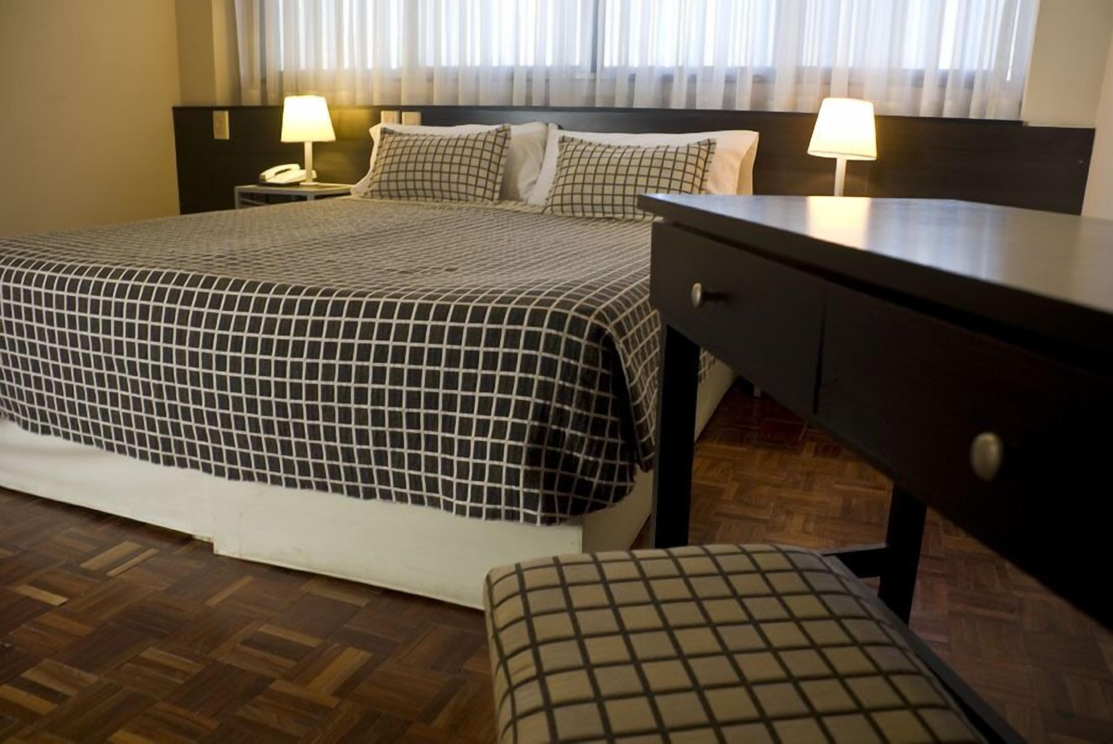 Habitación de hotel con una cama grande, dos mesitas de noche con lámparas, suelo de madera y vistas al exterior a través de cortinas translúcidas.