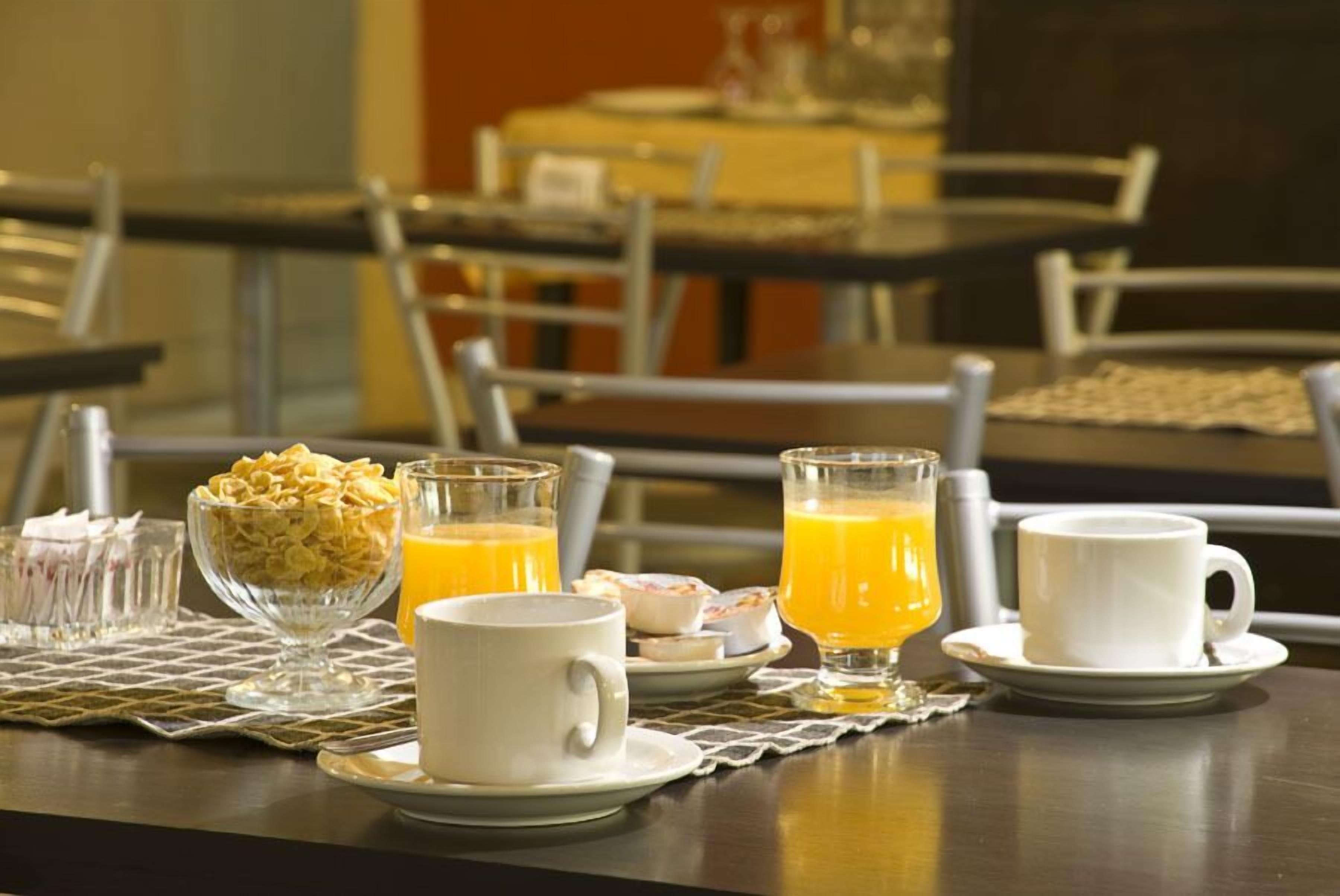 Desayuno con cereales, jugo de naranja y tazas de café sobre una mesa.