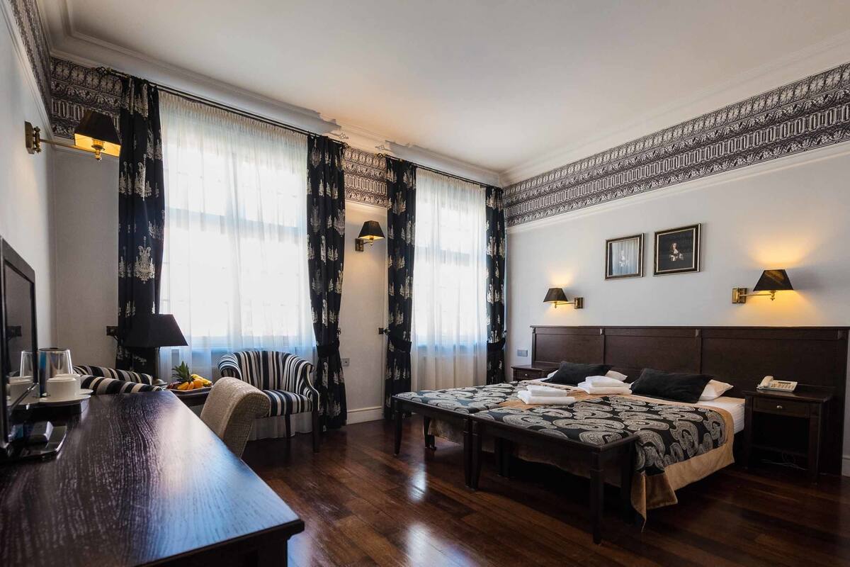 Una habitación de hotel con cama, mesita de noche, lámpara, silla y escritorio con televisor y tazas.