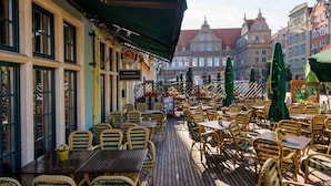 Een terras met rieten stoelen en tafels, een vlag en uitzicht op historische gebouwen op de achtergrond.