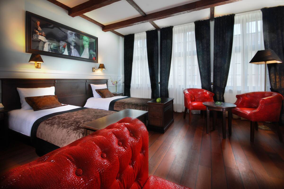 Habitación de hotel con dos camas, un sillón rojo, una mesita y un cuadro en la pared.