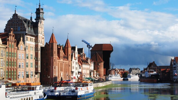 Een historisch stadsbeeld met een rivier, inclusief een opvallend gebouw met een toren en een groot schip afgemeerd aan de waterkant.
