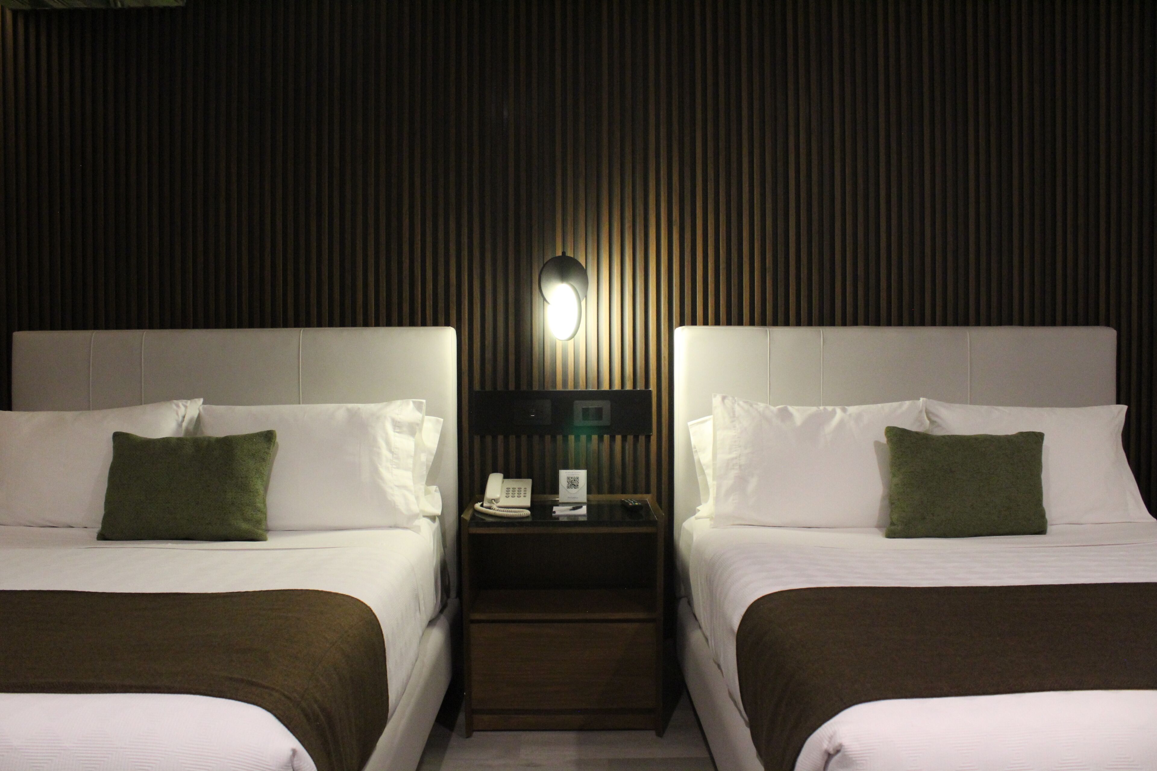 A hotel room with two beds, a nightstand with a phone and a lamp, and a wall-mounted light fixture.