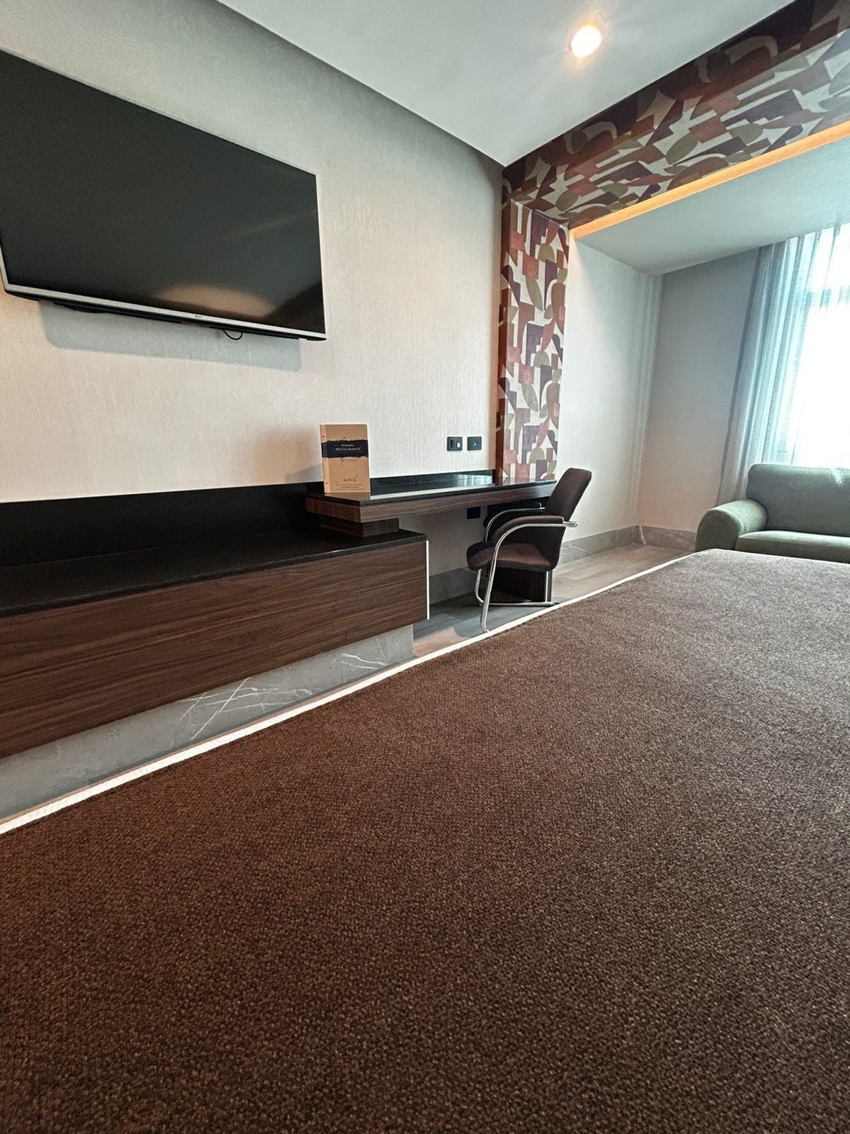 A modern hotel room with a flat-screen TV mounted on the wall, a desk with a chair, and a large window providing natural light.