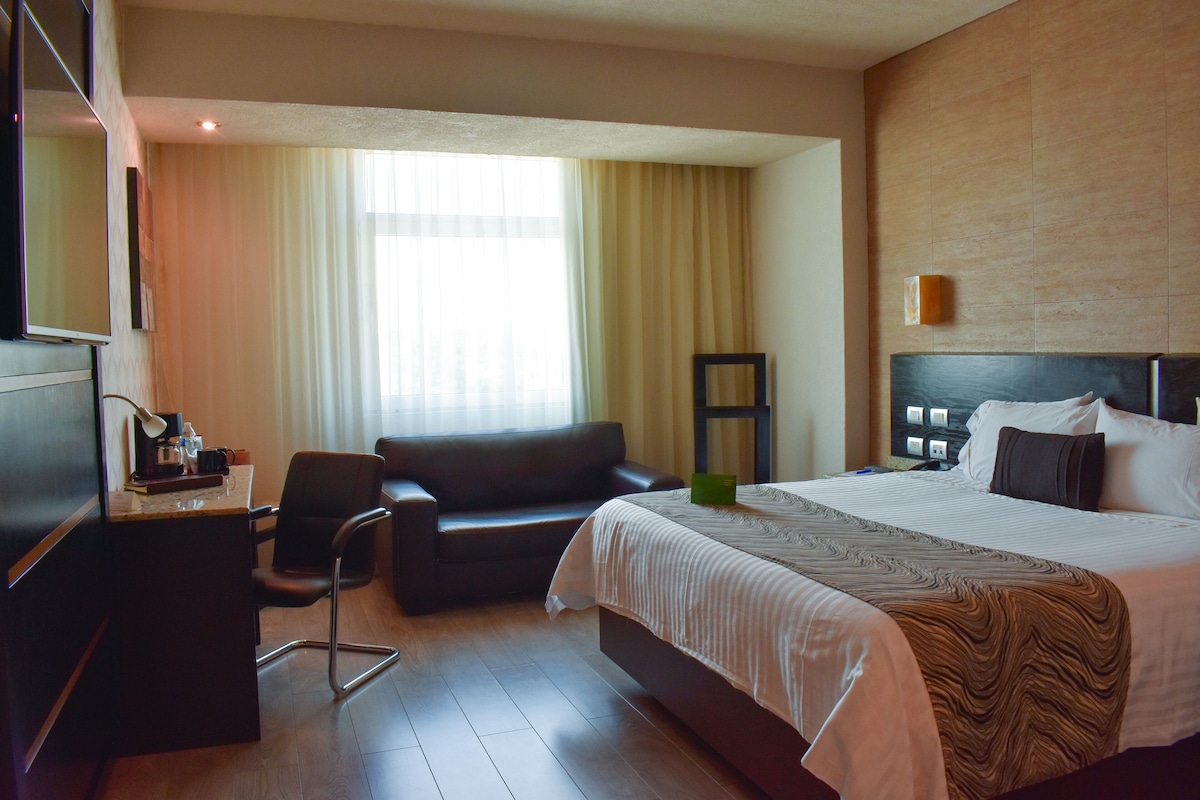 A hotel room with a bed, a desk, a chair, a sofa, and a window with curtains.