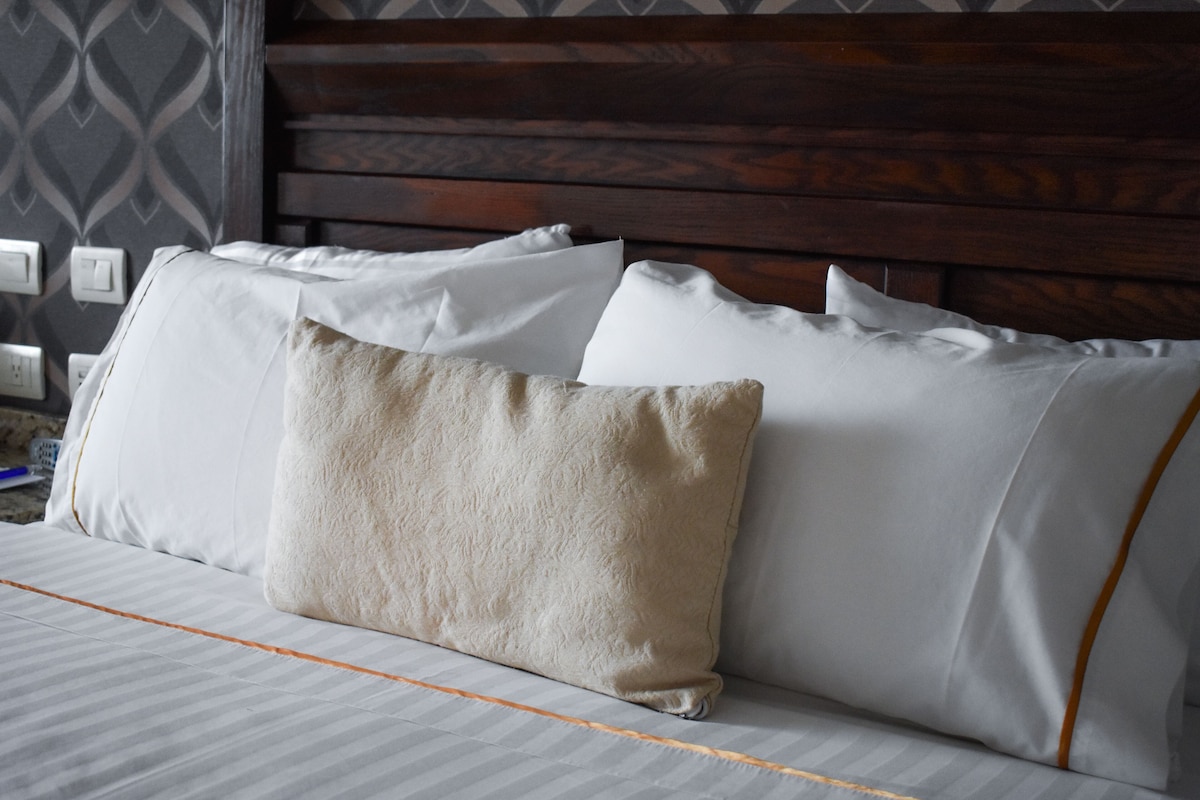 A bed with white linens and a beige pillow, a wooden headboard, and a patterned wall.