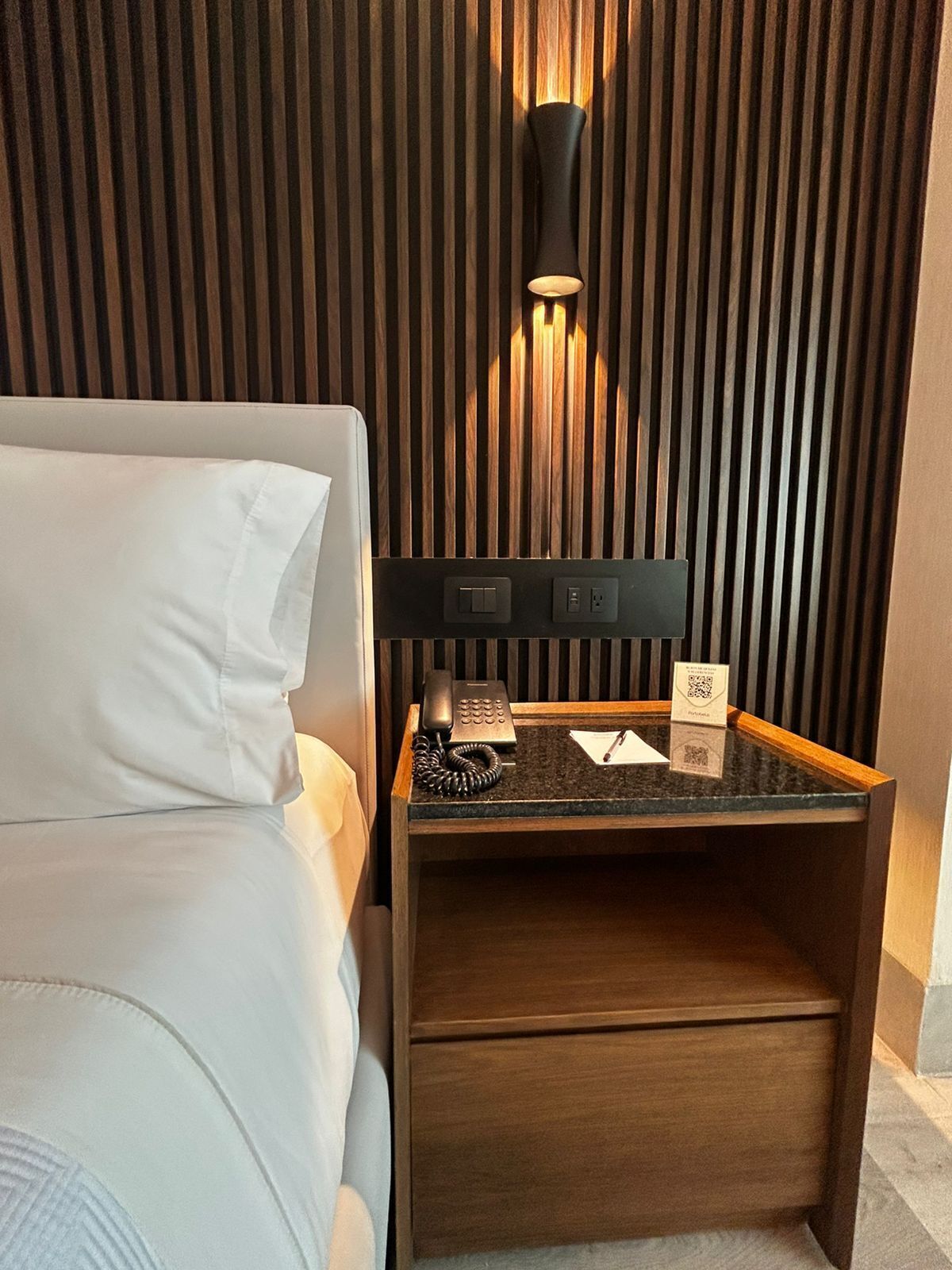 Een hotelkamer met een bed, een nachtkastje met telefoon, lamp en een klein lade.