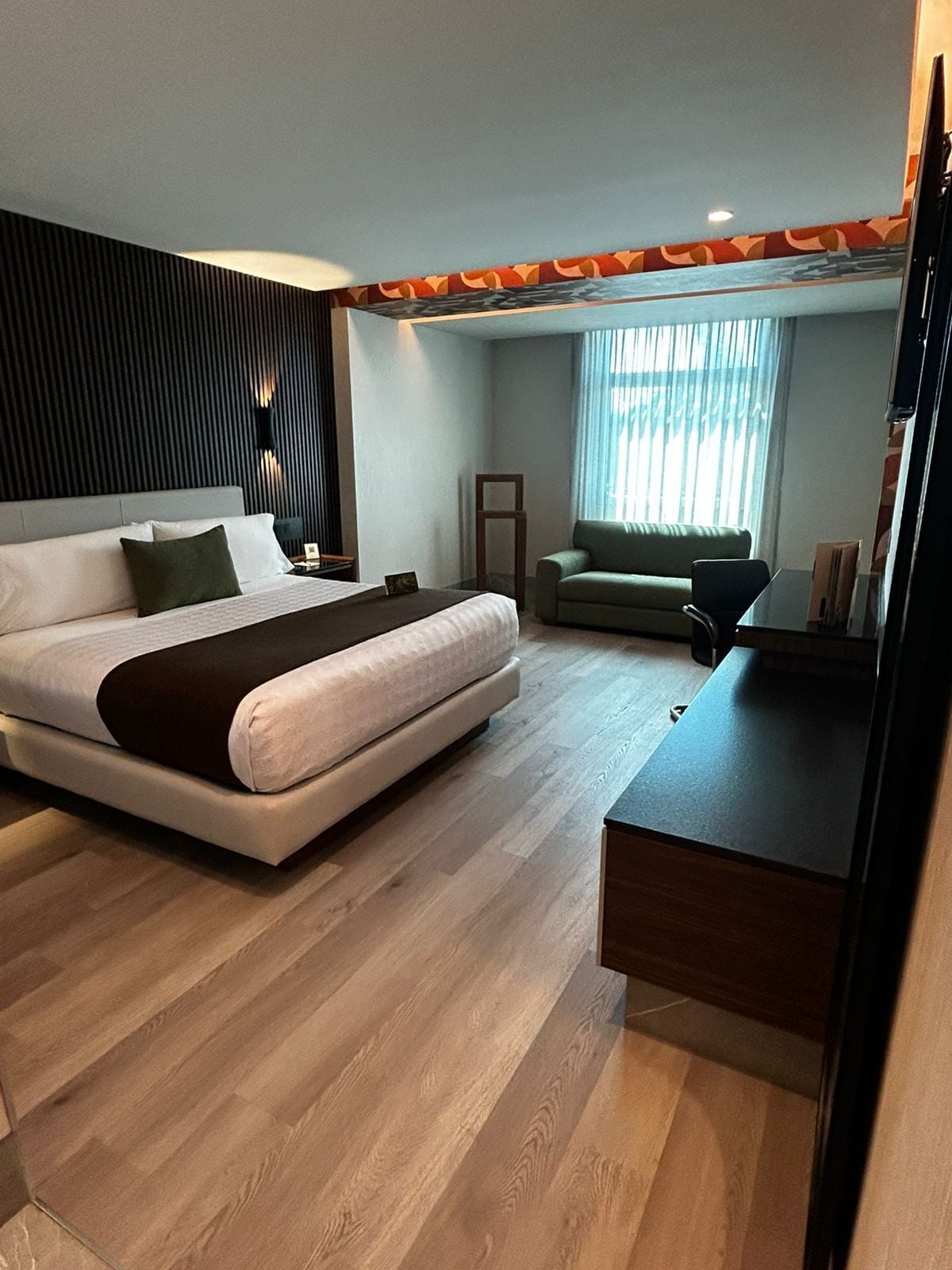 A modern hotel room with a large bed, a desk, a chair, and a window with blinds.