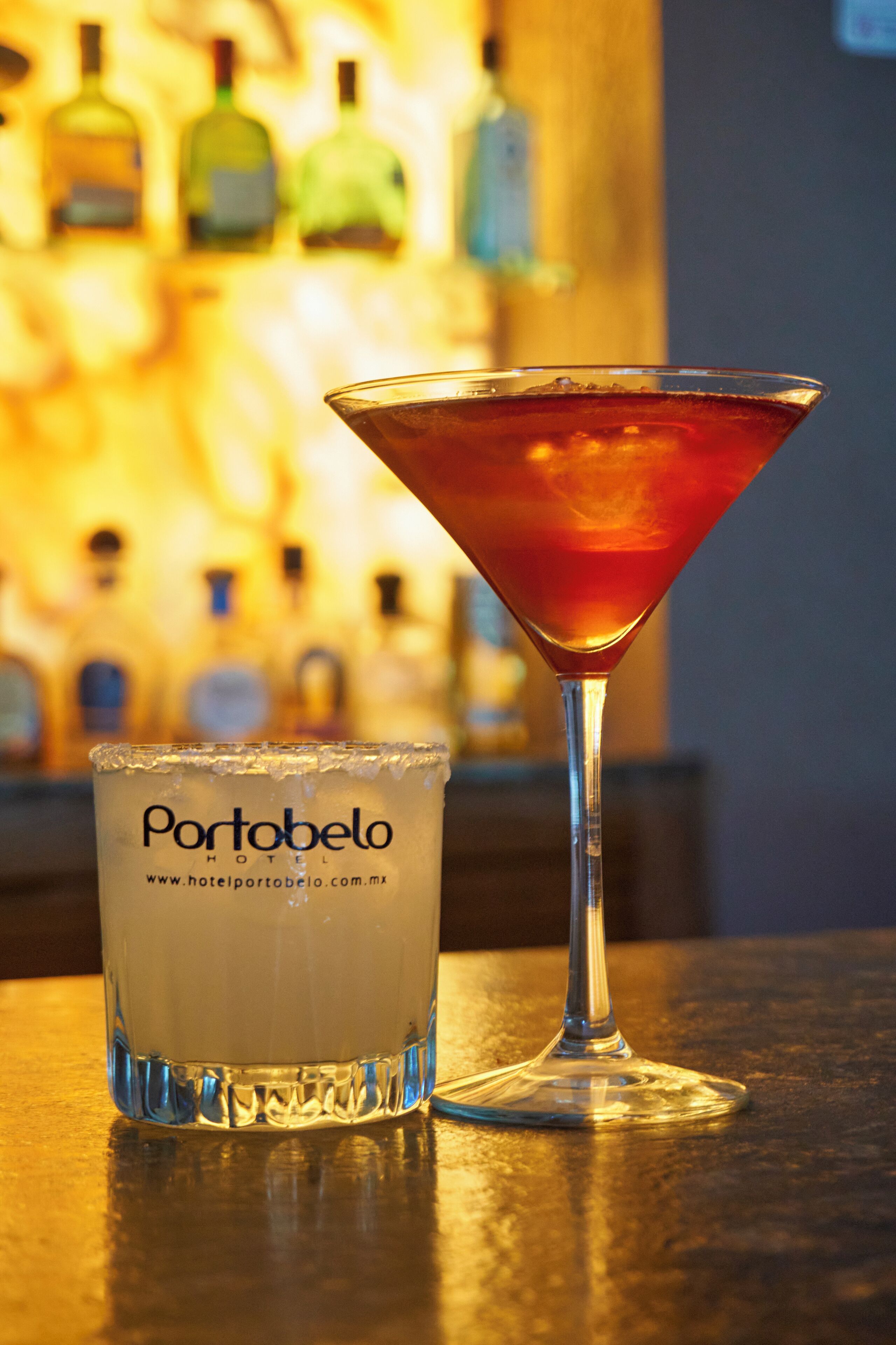 A martini glass and a glass of white beverage with the text "Portobelo HOTEL" on it, placed on a bar counter with a blurred background of bottles.