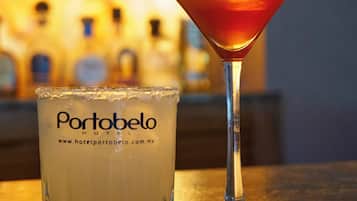 A martini glass and a glass of white beverage with the text "Portobelo HOTEL" on it, placed on a bar counter with a blurred background of bottles.