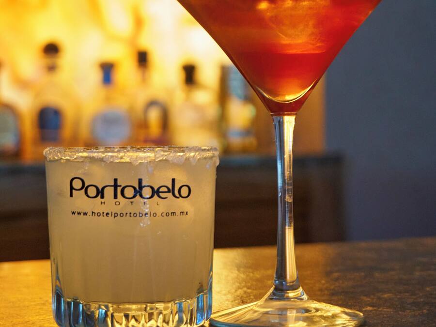 A martini glass and a glass of white beverage with the text "Portobelo HOTEL" on it, placed on a bar counter with a blurred background of bottles.