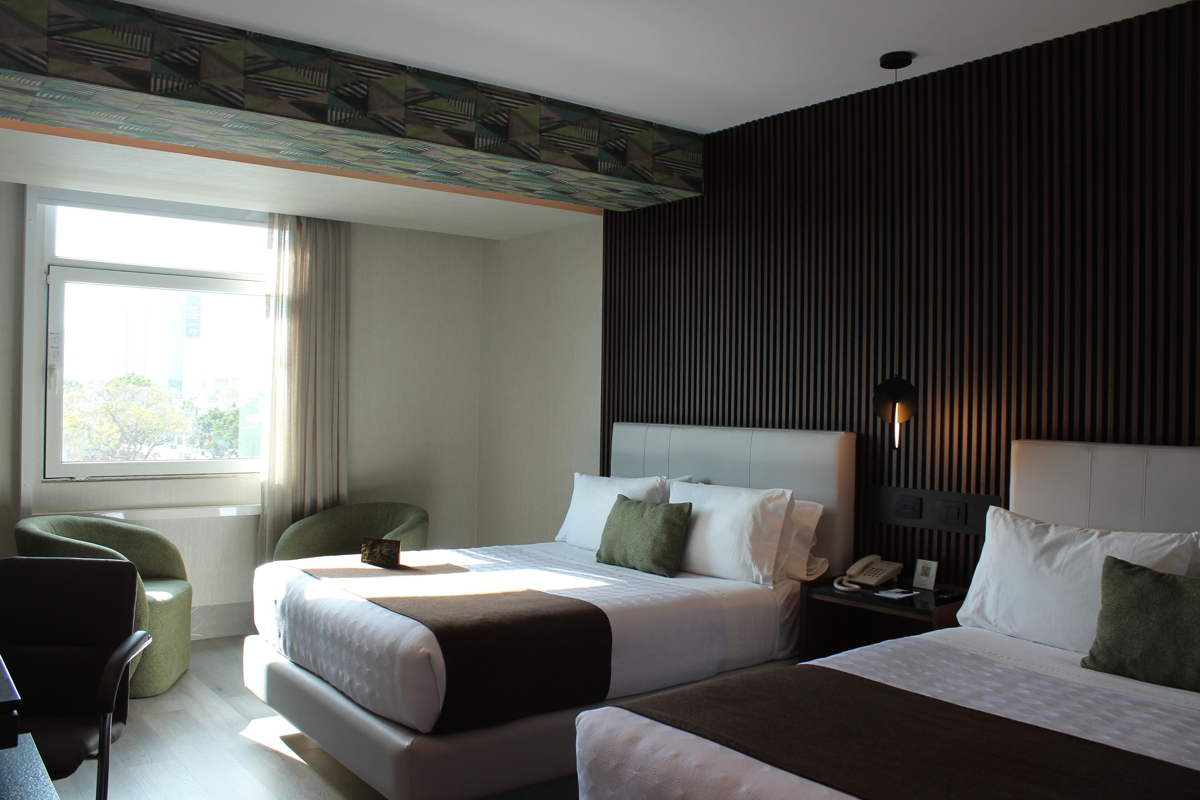 A hotel room with two beds, a desk, a chair, a window, and a wall with vertical lines.