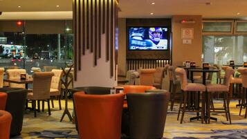 Um saguão de hotel com várias áreas de estar, uma tela de televisão e vista para a cidade através de grandes janelas.