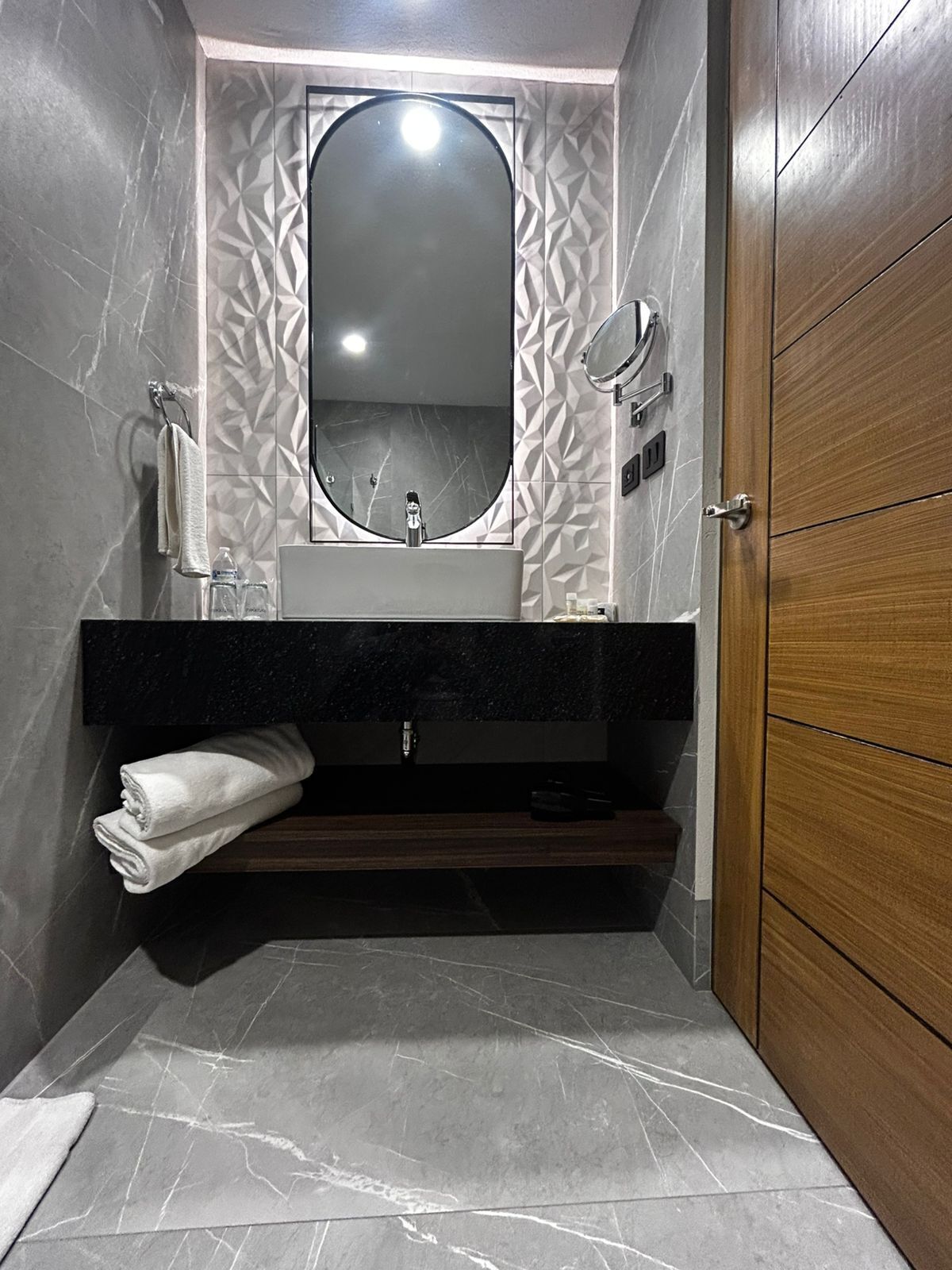 Un baño con suelo de mármol, puerta de madera y espejo sobre el lavabo.