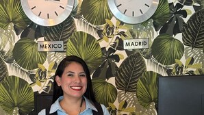 Una recepción con un reloj que muestra las etiquetas 'MEXICO' y 'MADRID', un monitor de computadora y un panel de madera agrietado.