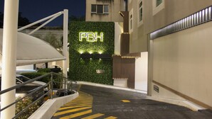 Entrada de hotel con pared verde y letrero iluminado 'PBH', zona de estacionamiento techada y una escalera que lleva a la entrada.