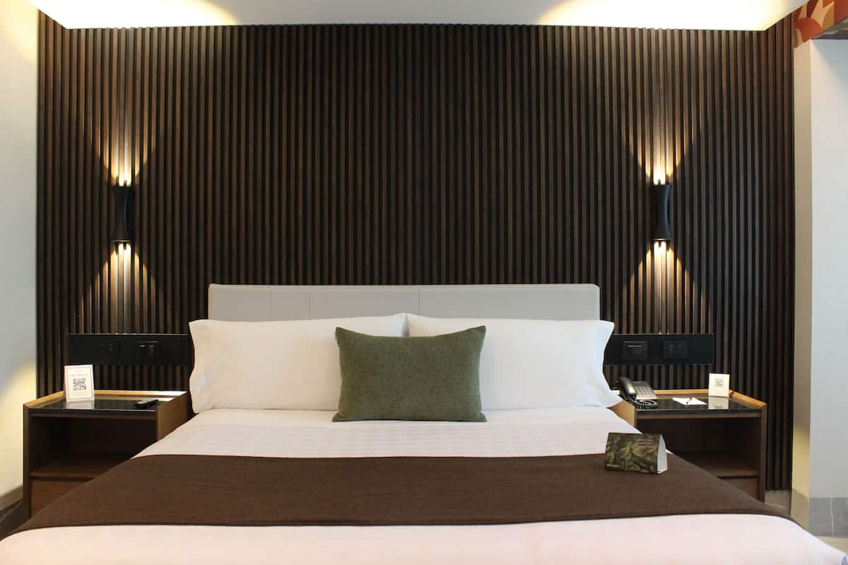 A hotel room with a large bed, two bedside tables, a nightstand, a pillow, a blanket, and a wall-mounted lamp.