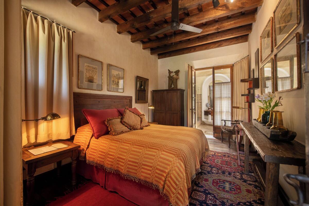A bedroom with a wooden ceiling, a bed with a patterned bedspread, a nightstand with a lamp, and a desk with a vase.