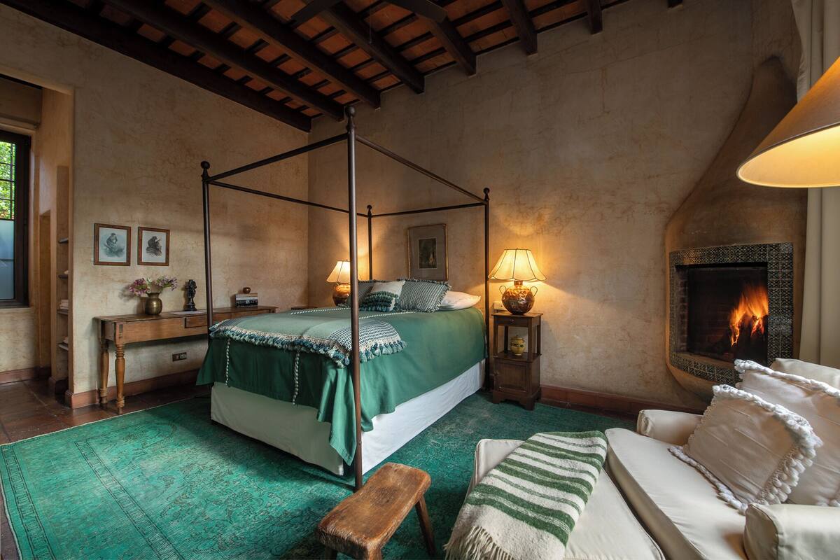 A four-poster bed with a green bedspread, a wooden bench, a white sofa, and a fireplace with a visible fire.