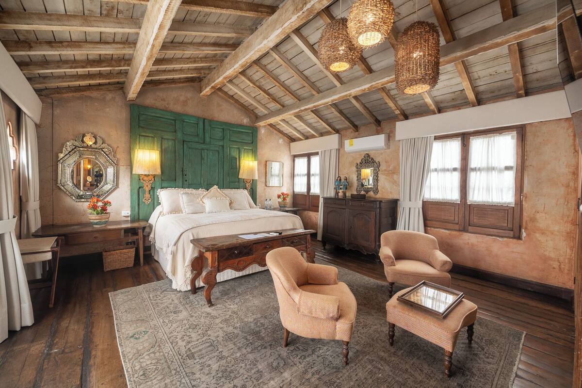 A spacious bedroom with a large bed, two armchairs, a wooden desk, and a dresser. The room features exposed wooden beams, a green door, and multiple windows with curtains.