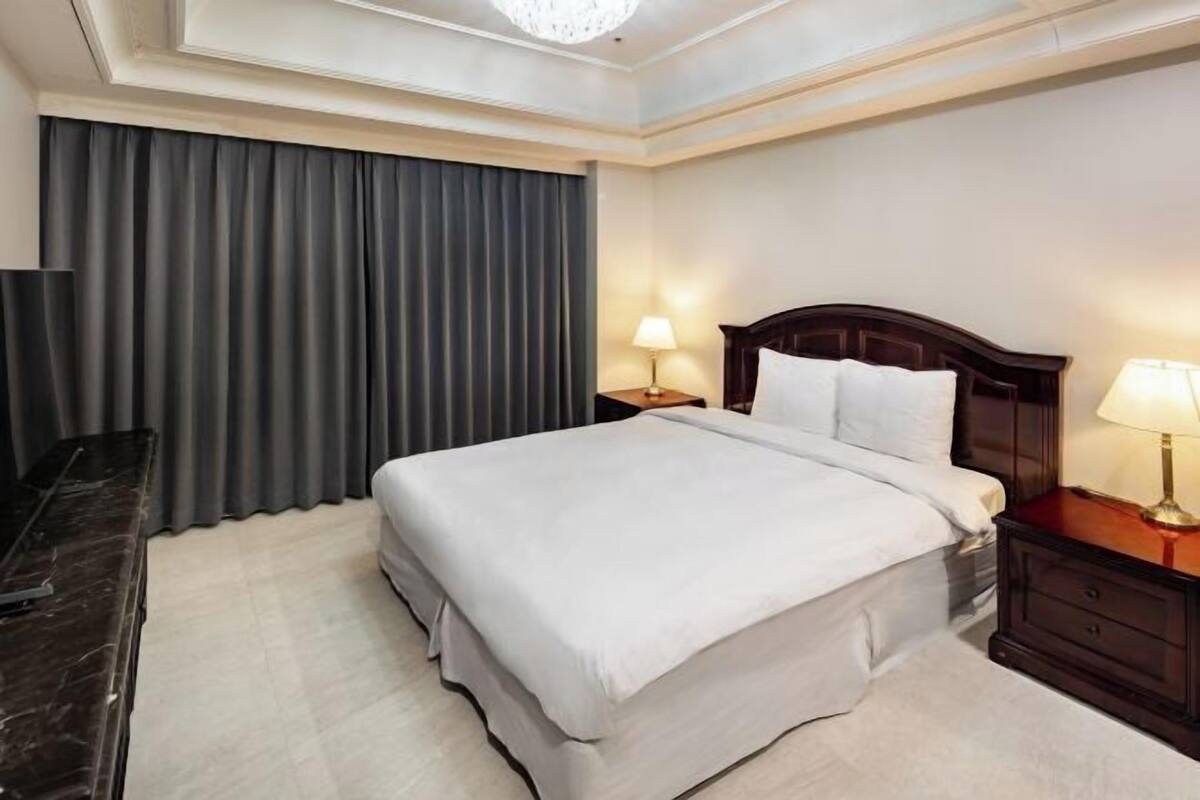 Ein Hotelzimmer mit einem großen Bett, zwei Nachttischen mit Lampen, einem dunklen Holzkopfende und einem großen Fenster mit Vorhängen.