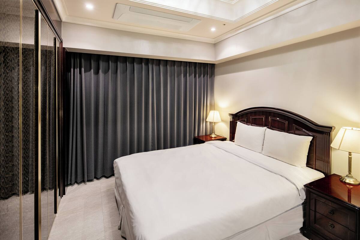 Ein Hotelzimmer mit einem großen Bett, einem Nachttisch mit Lampe, einem Spiegel und einem dunklen Vorhang.