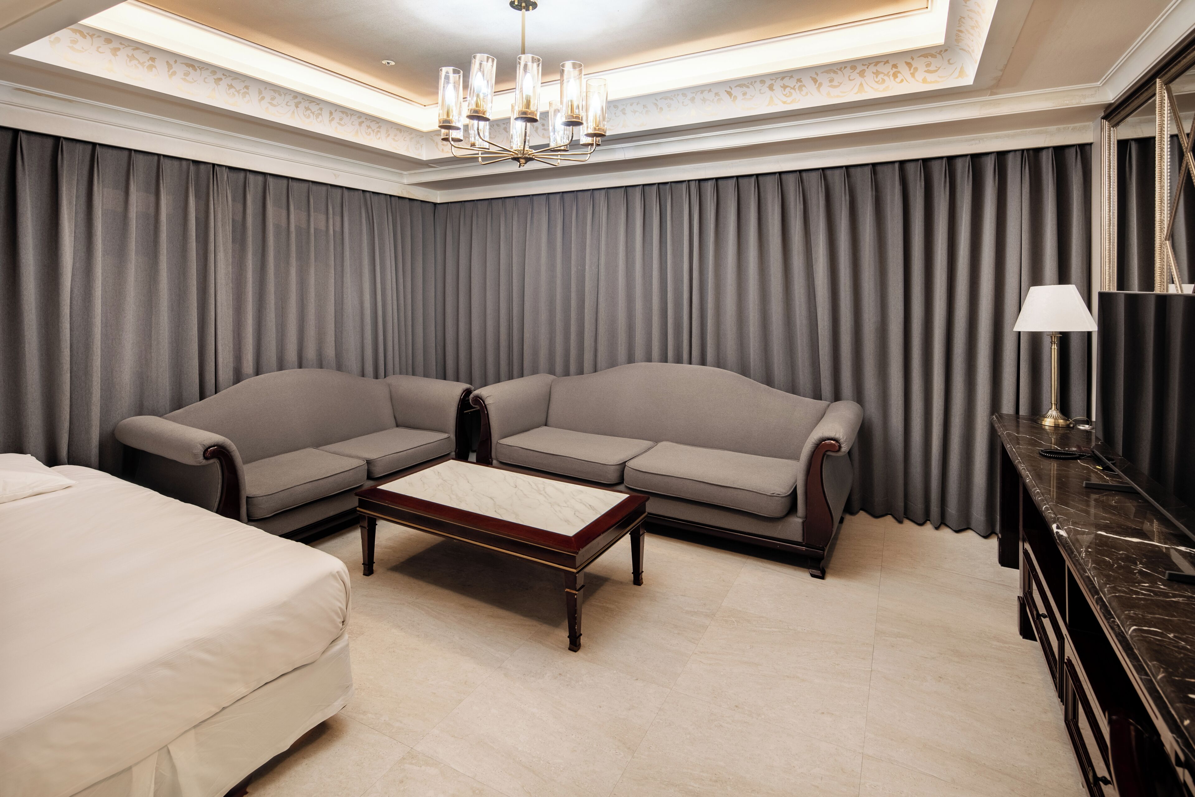 A hotel room with a large sofa, a smaller sofa, a bed, a coffee table, a desk, and a chandelier.