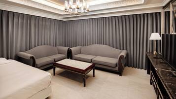 A hotel room with a large sofa, a smaller sofa, a bed, a coffee table, a desk, and a chandelier.