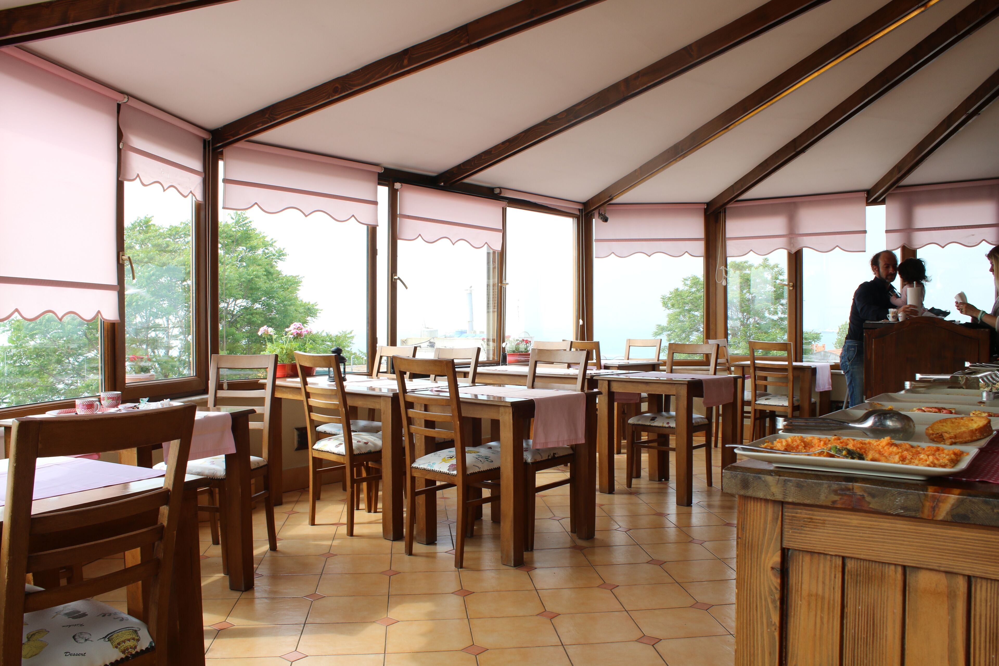 A dining area with wooden tables and chairs, large windows with pink curtains, and a buffet counter with food.