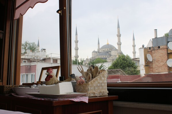 A view of a mosque with multiple minarets and a dome through a window, with a table in the foreground featuring a book, a picture frame, and a basket of wooden utensils. - Emine Sultan Hotel (Istanbul)