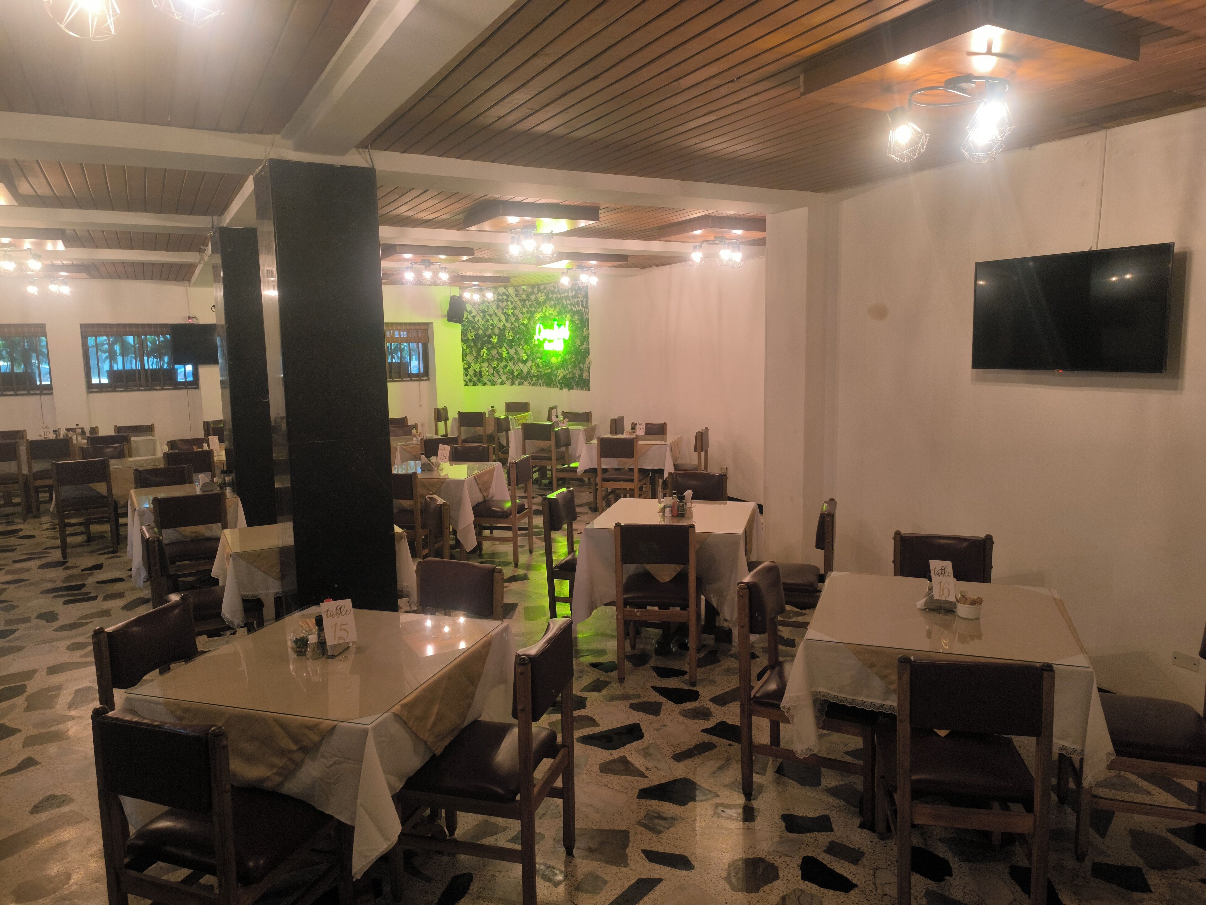 A restaurant with multiple tables and chairs, a green wall with a plant, and a TV on the wall.