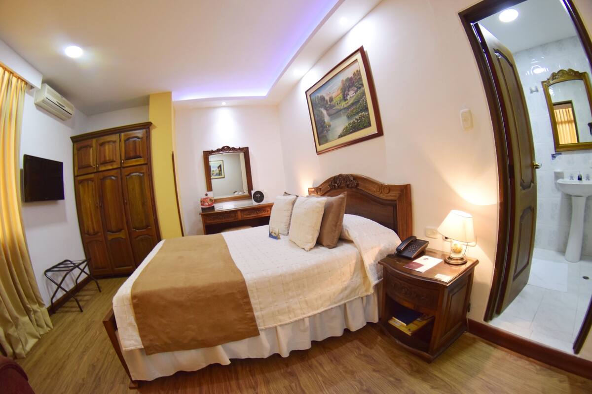 A hotel room with a bed, bedside table, lamp, mirror, wardrobe, and a painting on the wall.