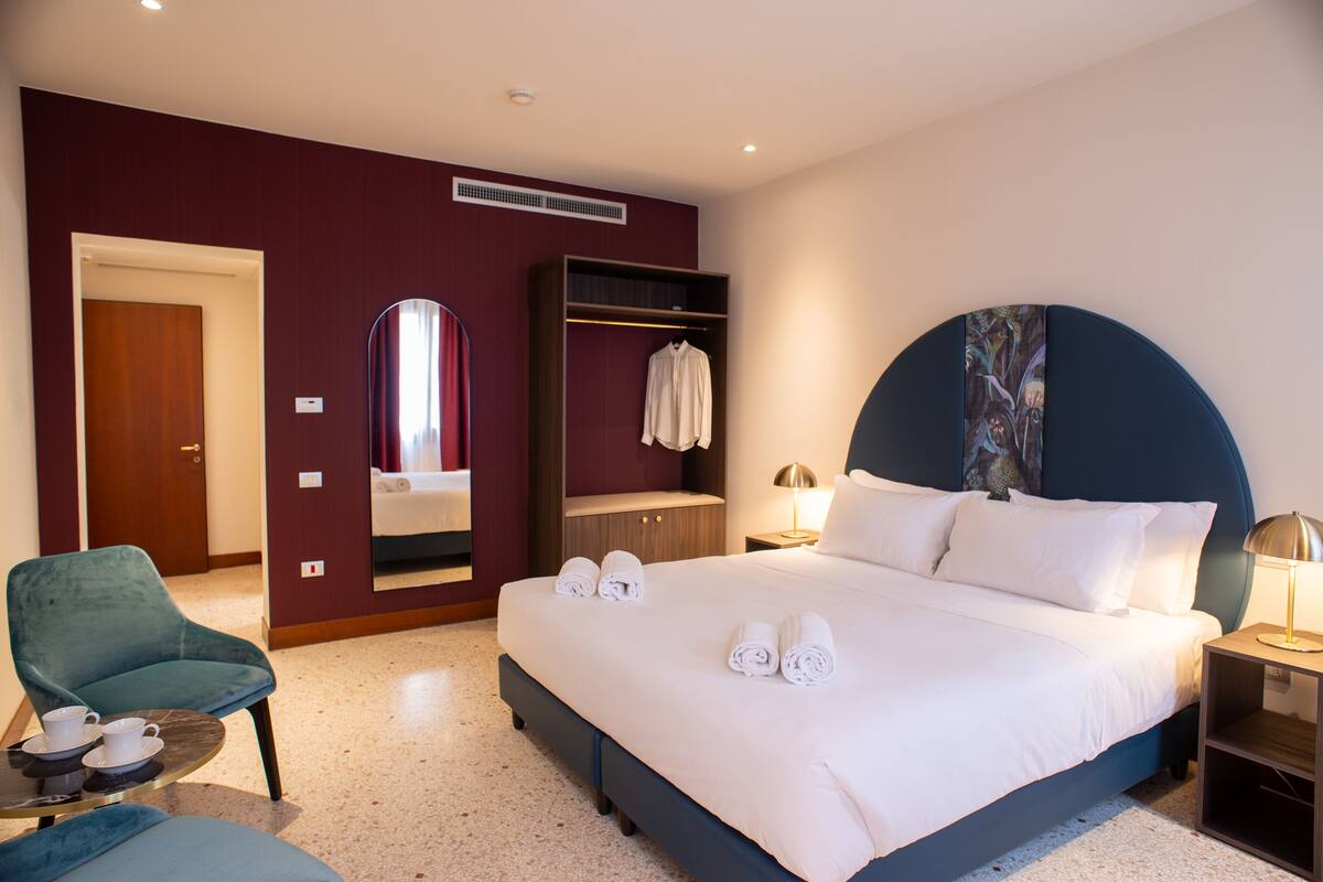 Habitación de hotel con una cama grande, zona de estar con una silla y una mesa, armario y espejo.