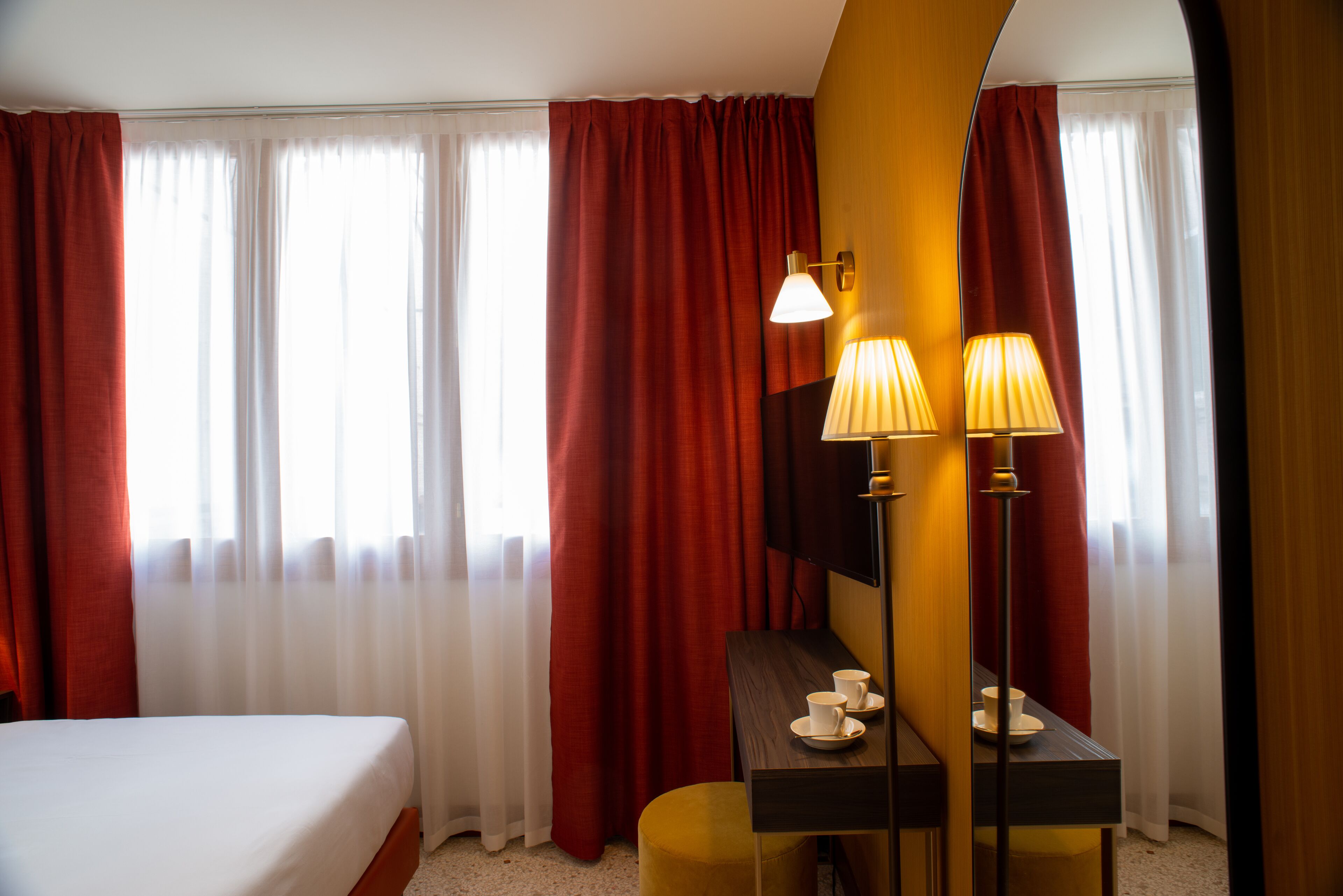 A hotel room with a bed, a desk with two lamps, a cup and saucer, and red curtains.