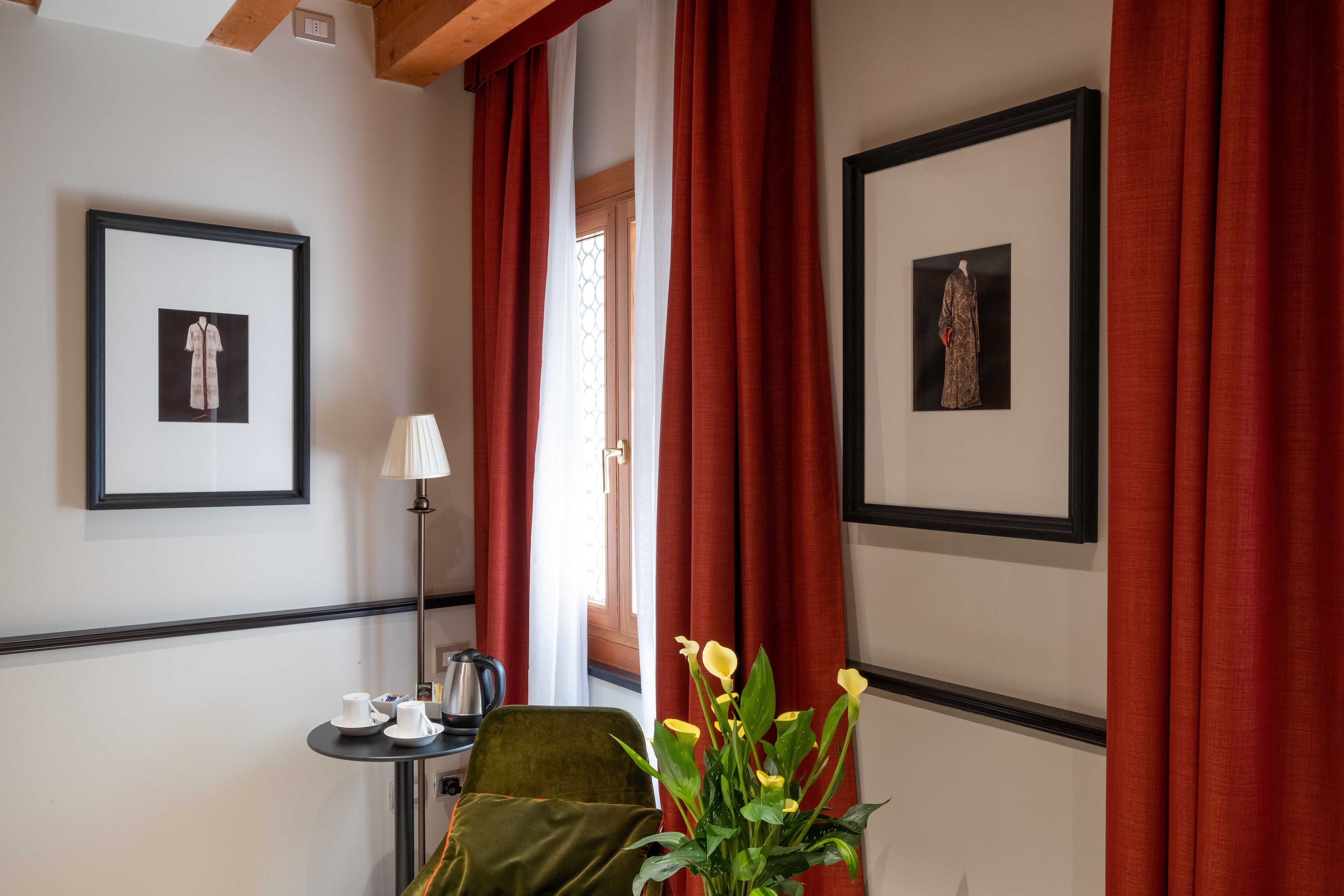 A hotel room with a green armchair, a small table with cups, a lamp, and two framed pictures on the wall.