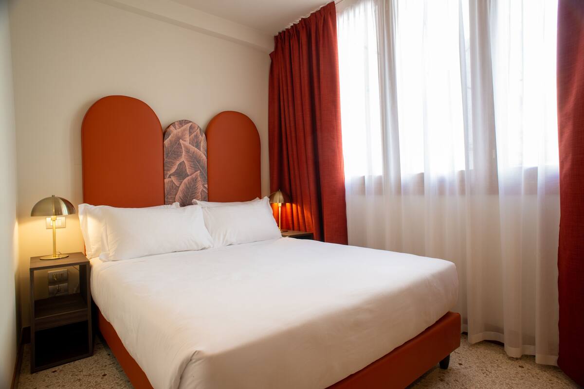 Una cama bien hecha con sábanas blancas, dos lámparas de noche y un ventanal con cortinas translúcidas.