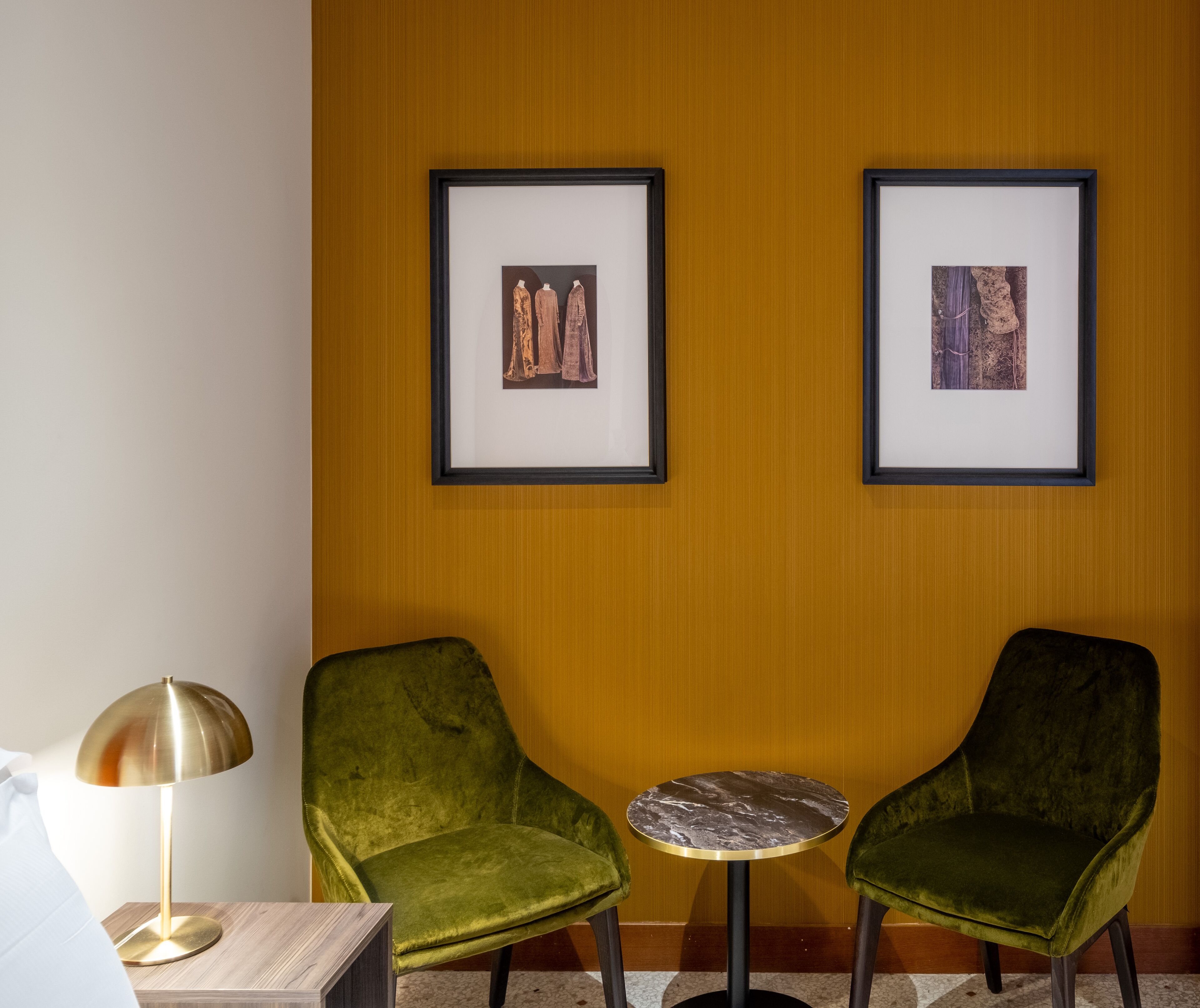 A room with two green velvet chairs, a small round table, and two framed artworks on the wall.