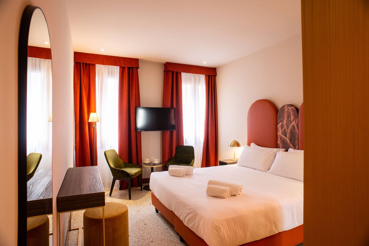 Habitación de hotel con cama, escritorio, silla, televisor y ventana con cortinas.