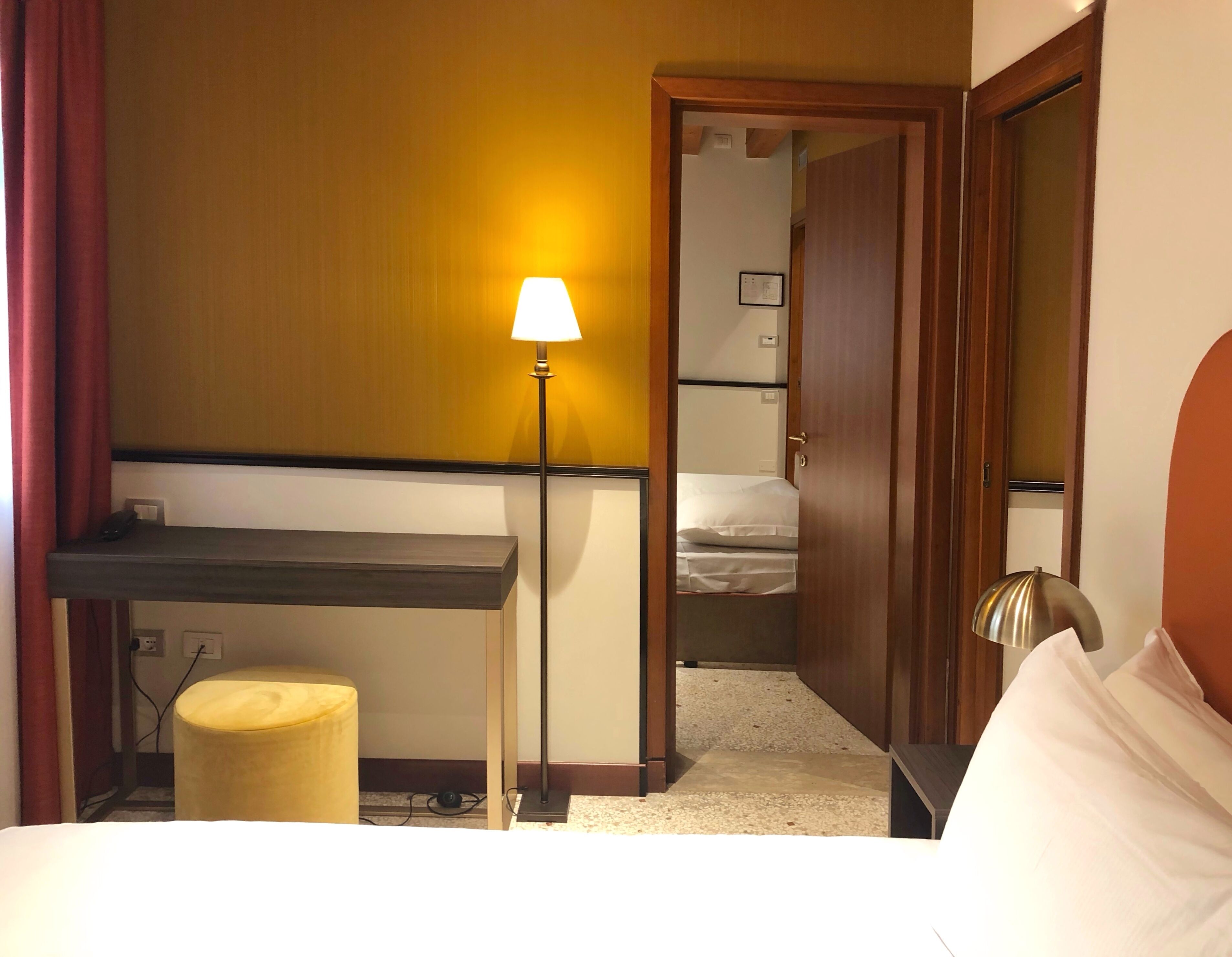 A hotel room with a bed, a desk, a lamp, and a door leading to a bathroom.