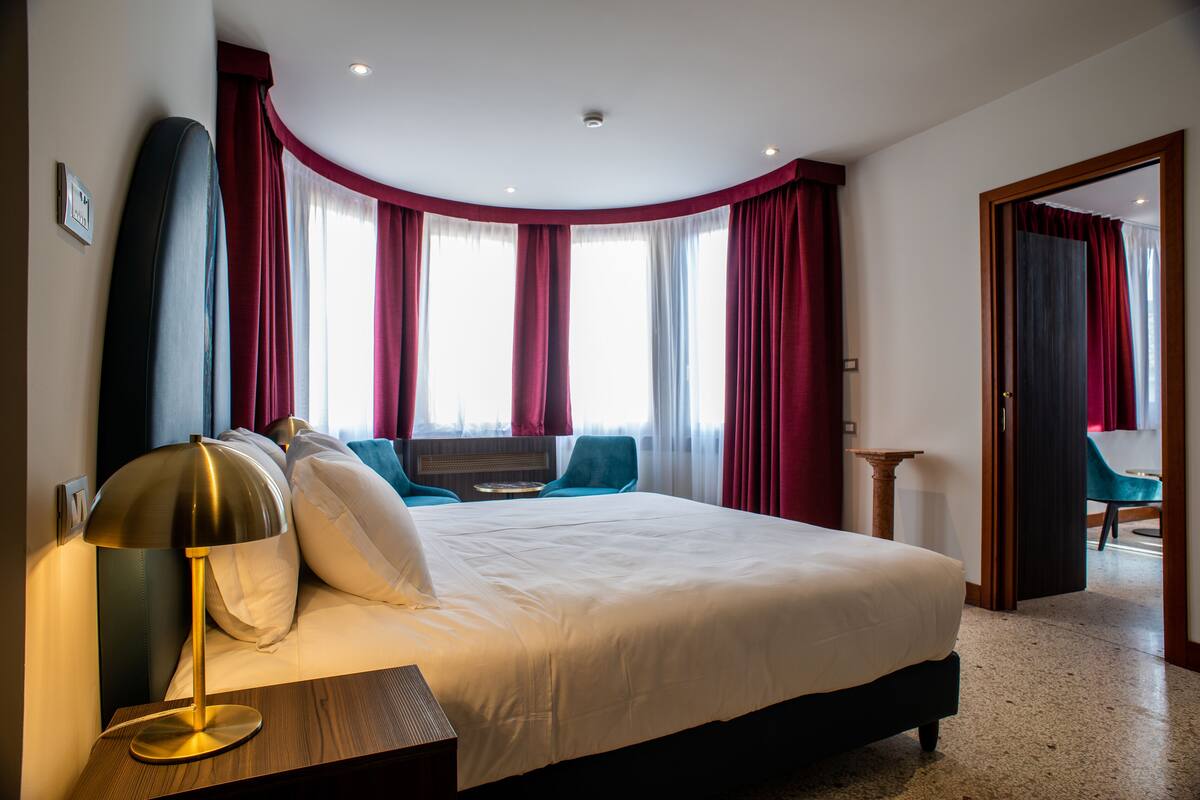 Una habitación de hotel con una cama grande, una mesita de noche con lámpara, una ventana con cortinas y vistas al exterior.