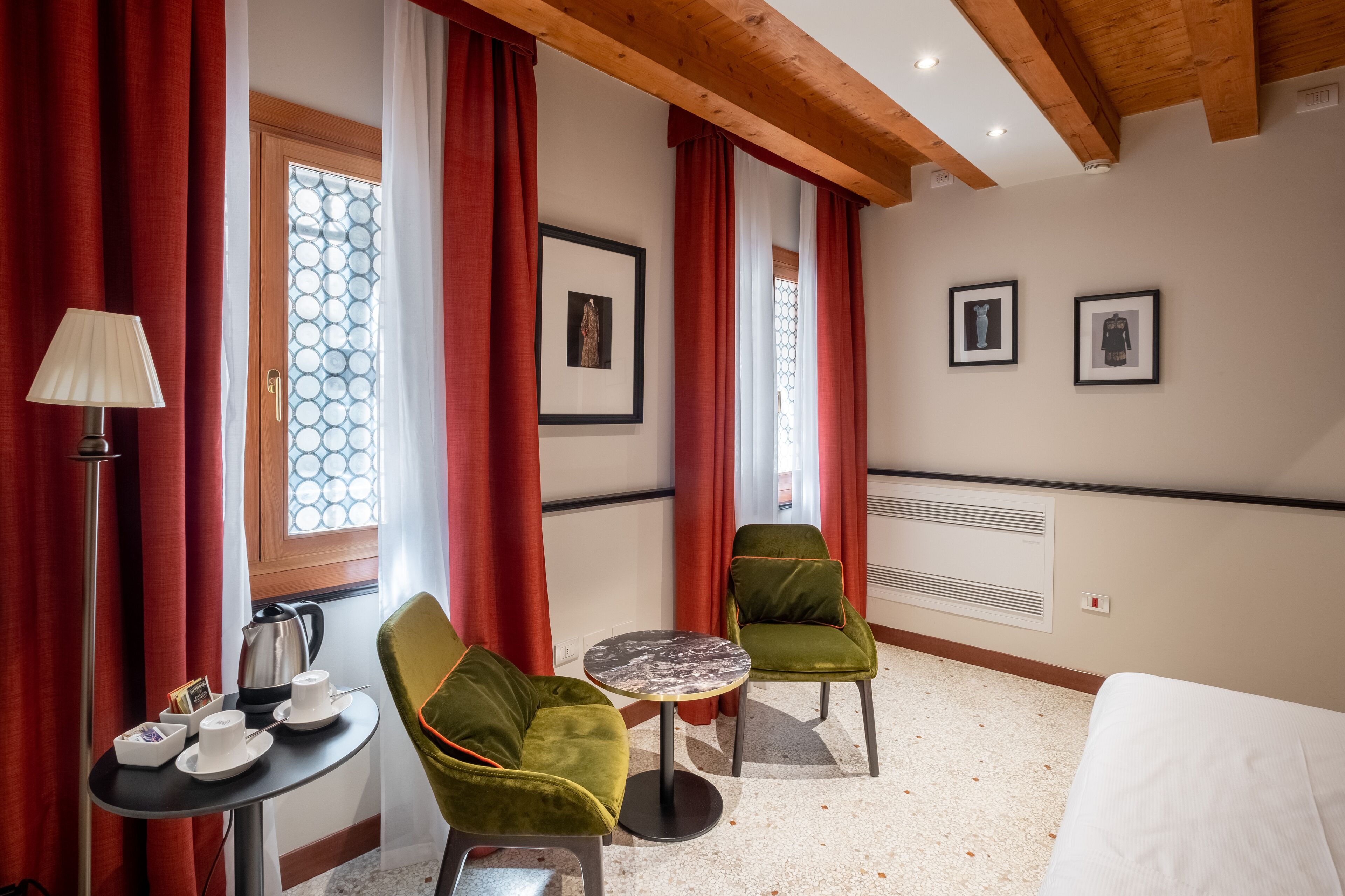 A hotel room with two green chairs, a small round table, a bed, and a window with red curtains.