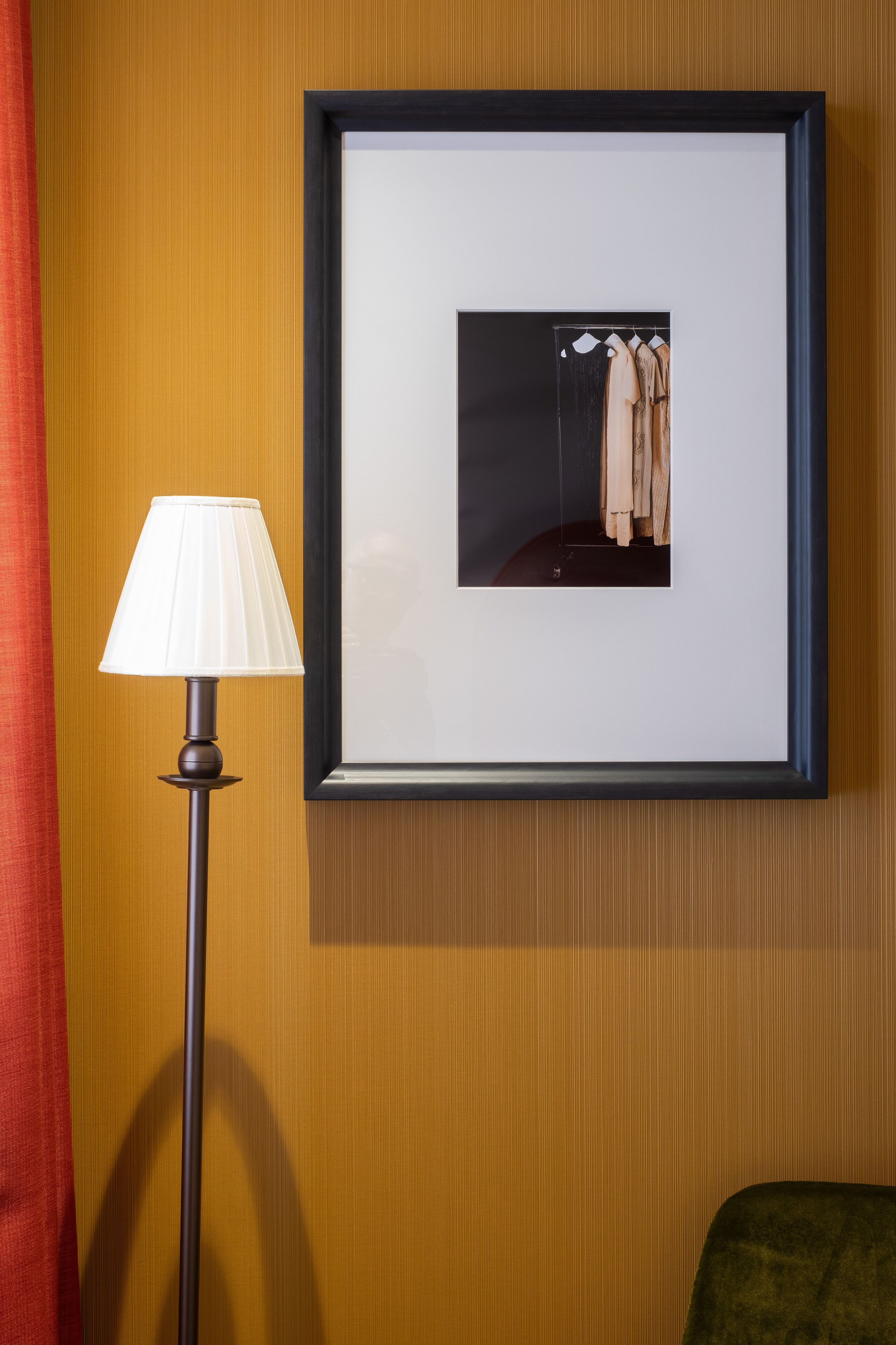 A framed photograph of a clothing rack on a wall, a floor lamp, and a red curtain.