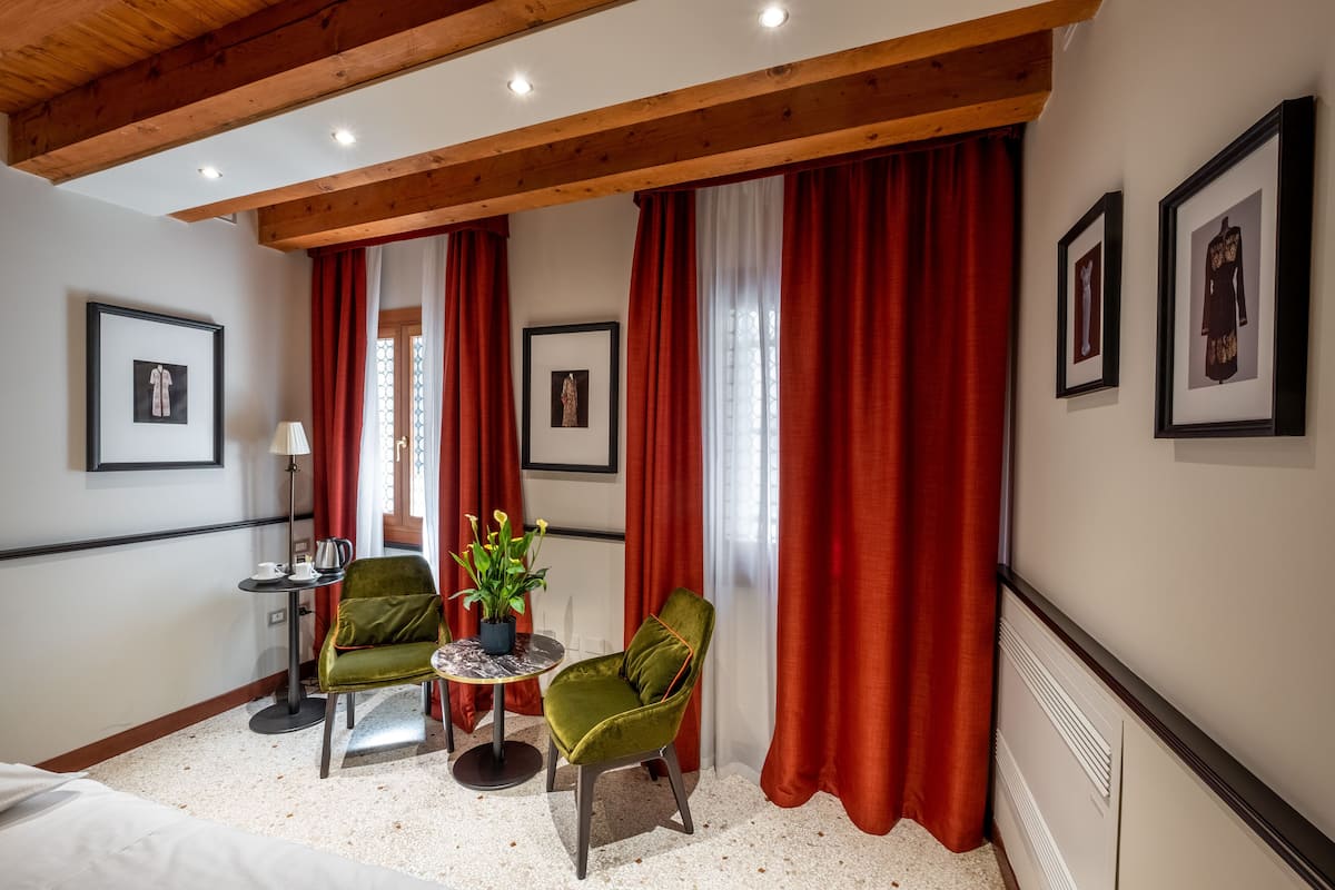 Habitación de hotel con una cama, dos sillas, una mesita con lámpara, una planta en maceta y cuadros en las paredes.