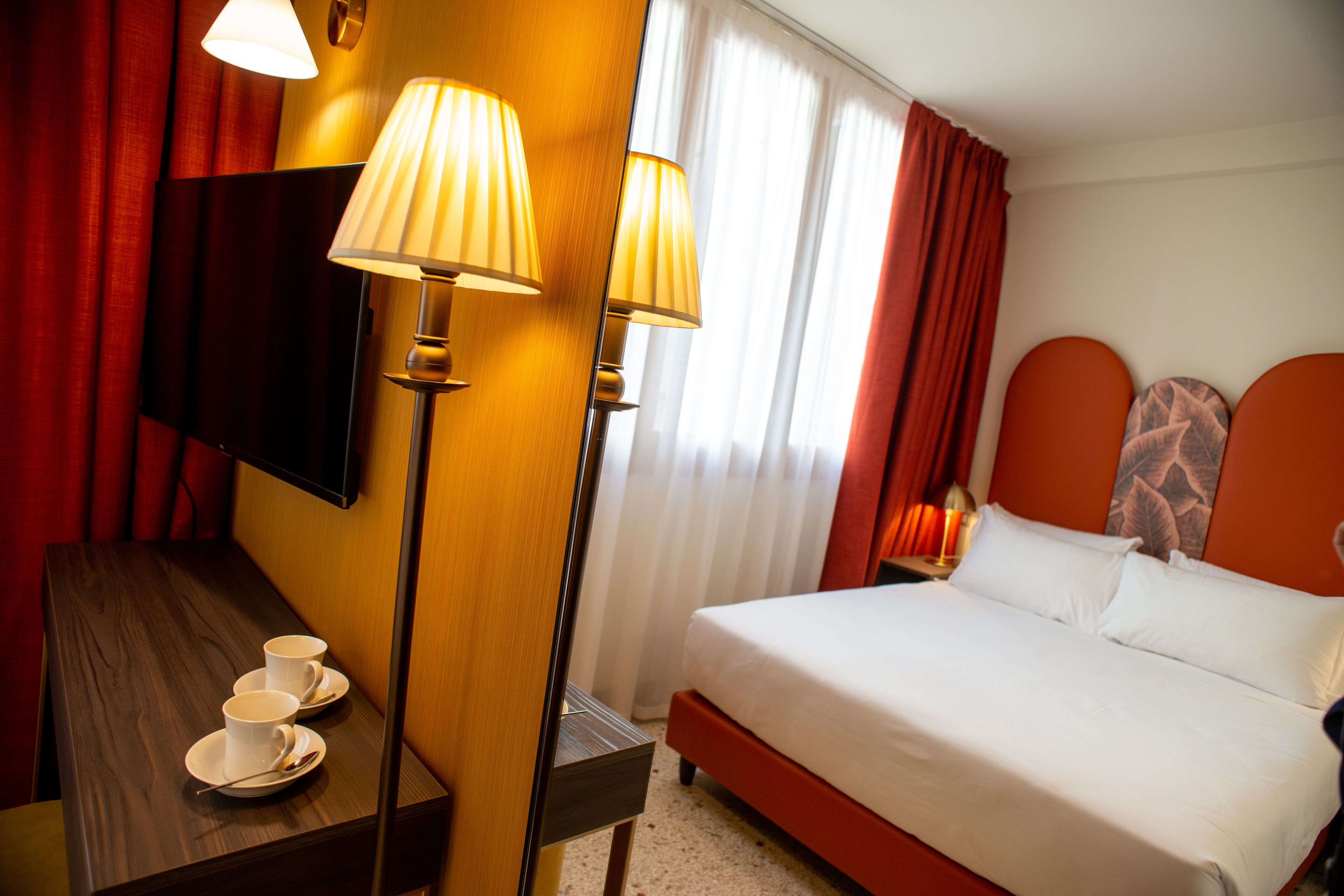 A hotel room with a bed, a desk with a lamp, a television, and a window with curtains.
