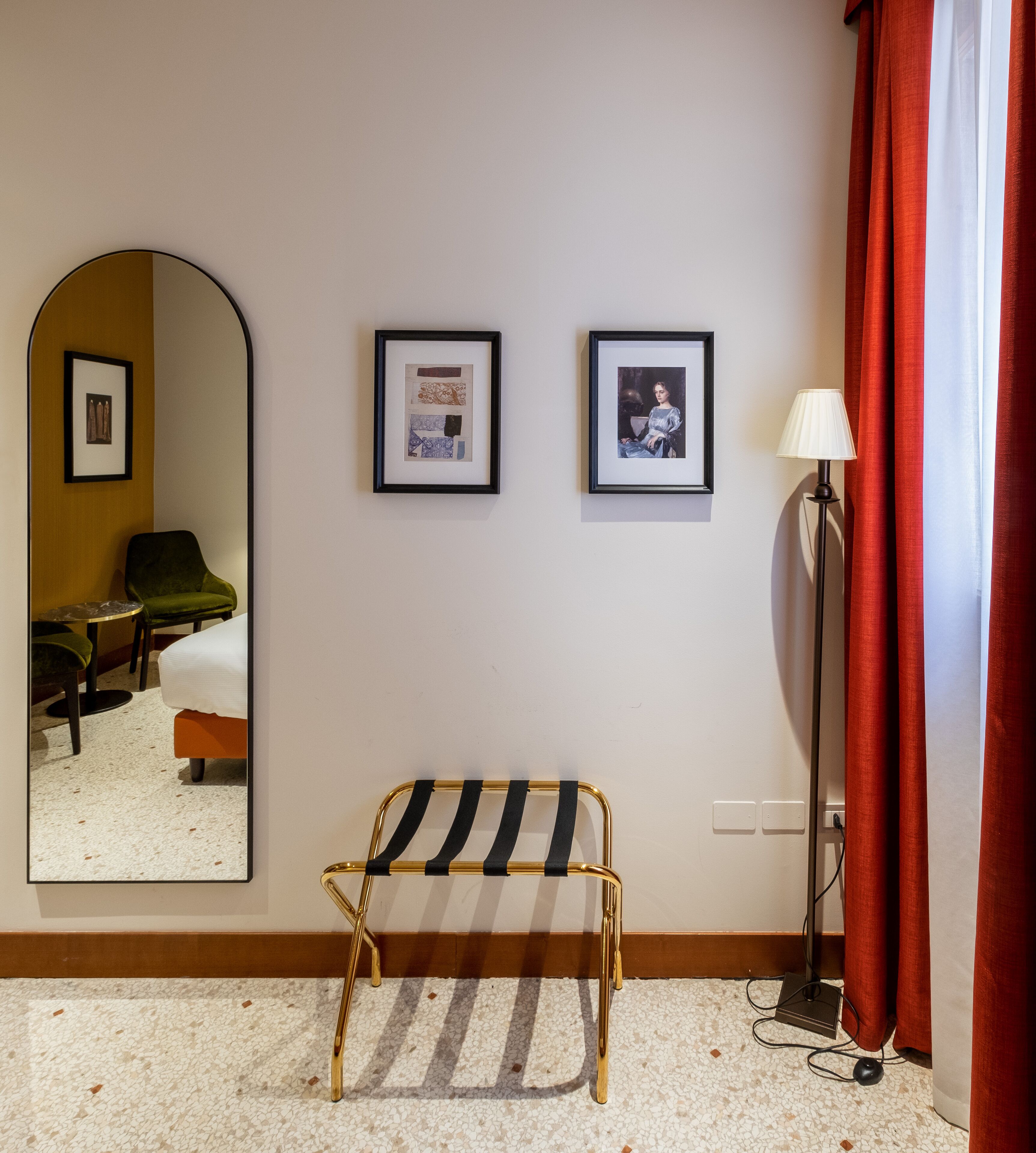 Habitació d'hotel amb un llit, una panxa, un mirall, dues fotografies amb marc, una làmpada i una cortina vermella.