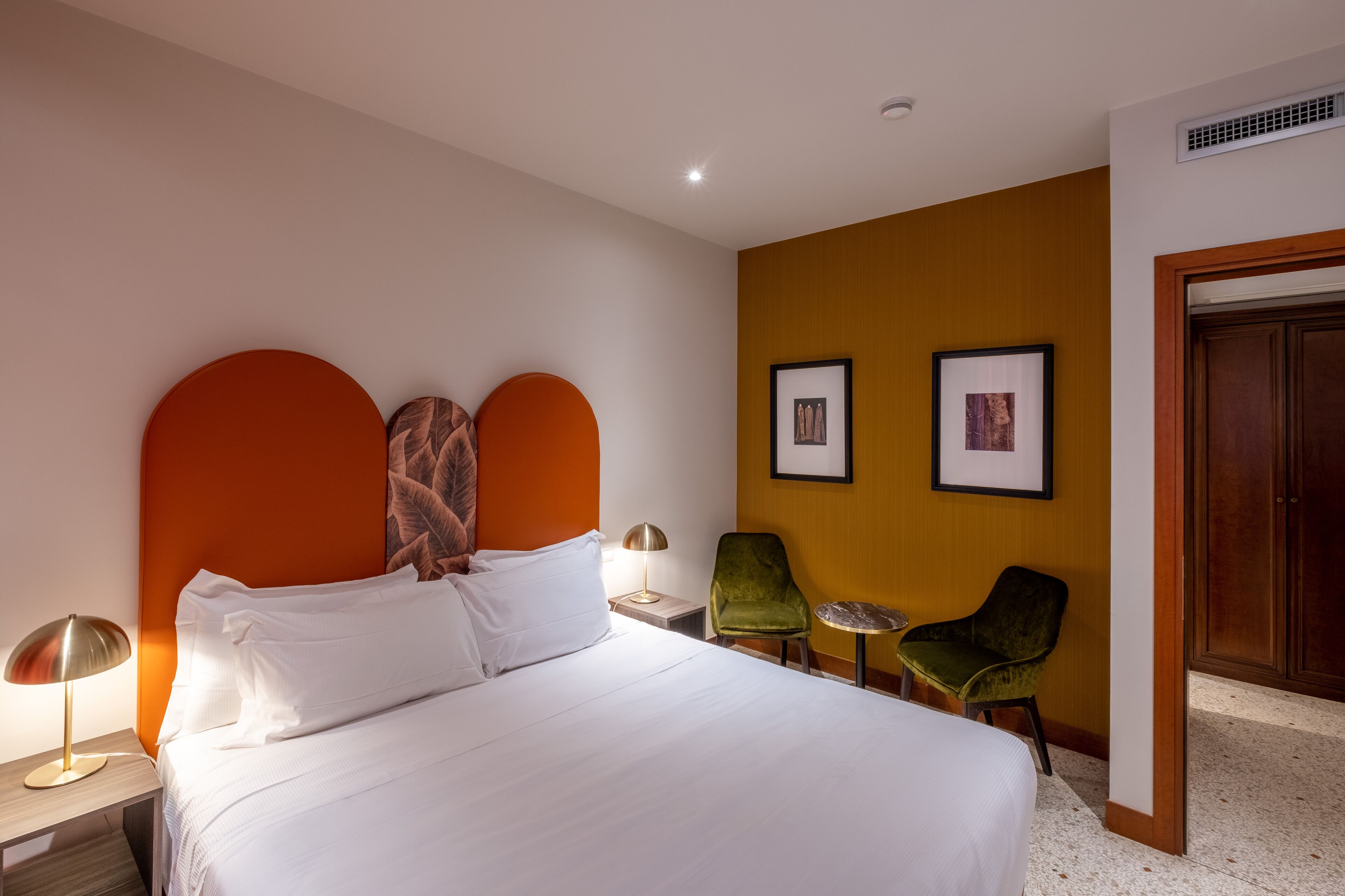 A hotel room with a large bed, two chairs, a small table, and framed pictures on the wall.